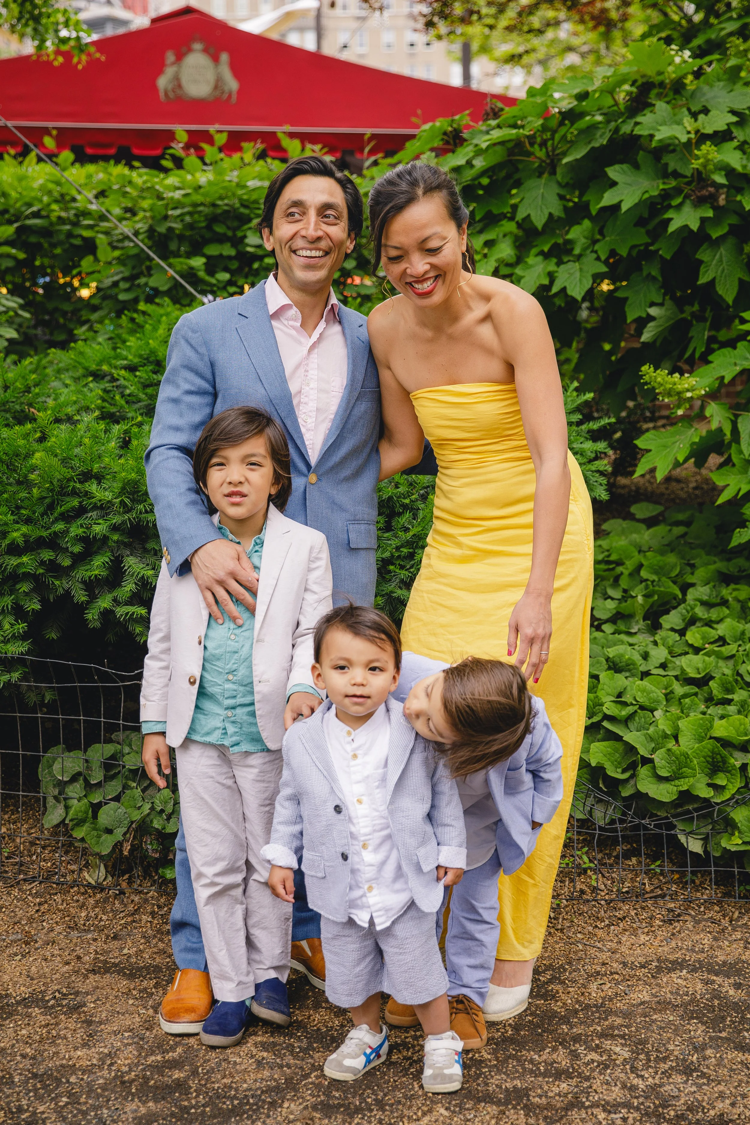 A group of six people, including three children and three adults, standing together outdoors in front of green leafy bushes. The adults are smiling and appear happy. The background includes a red canopy and buildings.