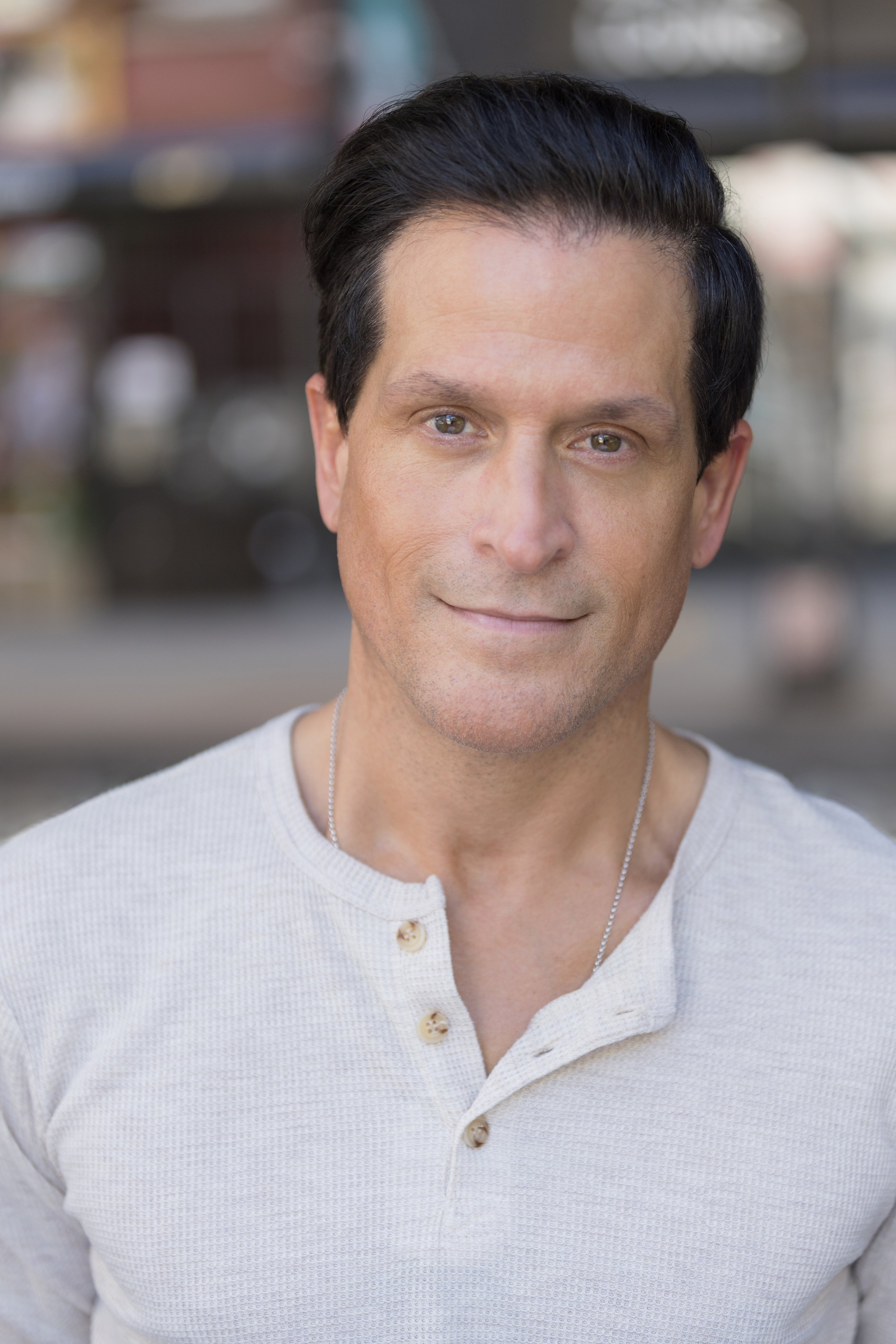 A close-up portrait of a man with dark, swept-back hair wearing a light beige button-up shirt with a silver necklace, outdoors with blurred city background.