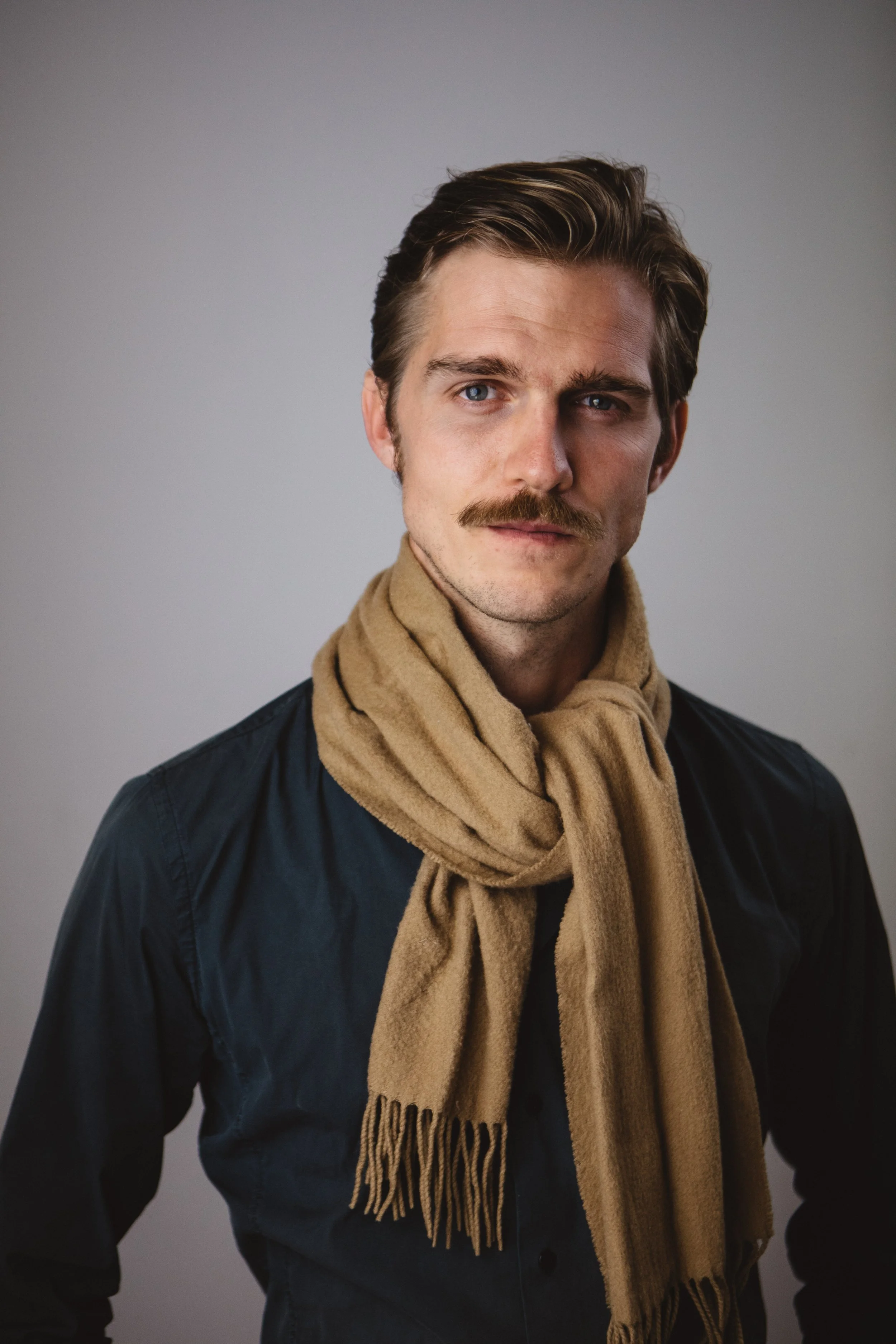 Portrait of a man with light skin, blue eyes, brown hair, wearing a beige scarf and a dark jacket against a plain gray background.