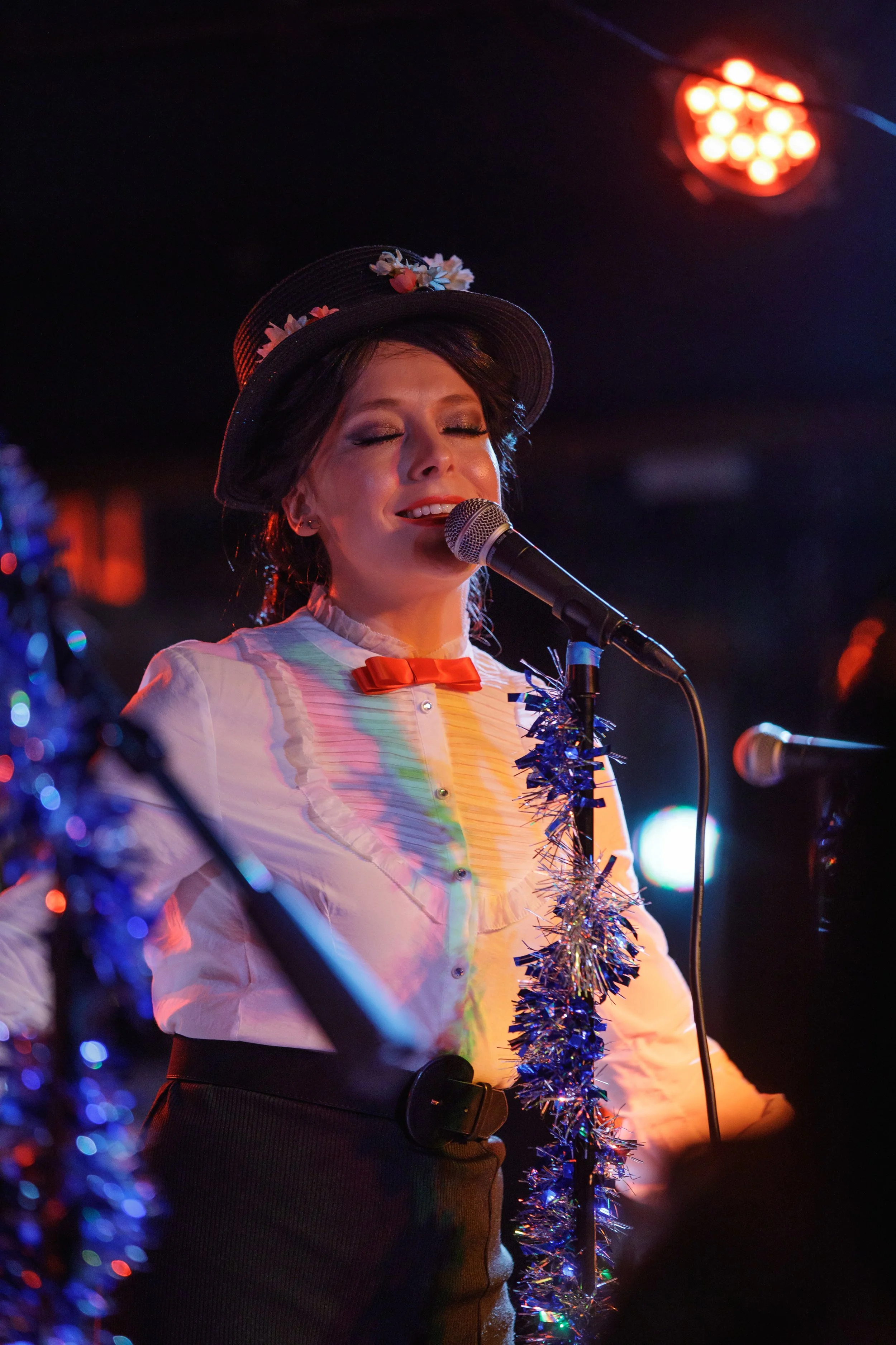 A woman singing into a microphone, wearing a black hat decorated with flowers, a white blouse with a red bow, and a tinsel garland around her neck, on stage with colorful lights.