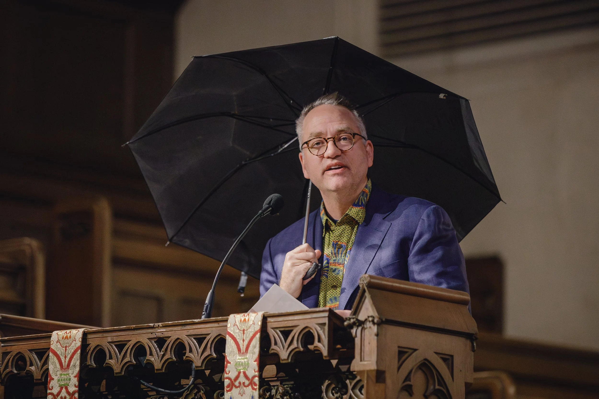 A man in a blue suit and patterned shirt is standing behind a wooden podium, holding a black umbrella over his head, speaking into a microphone.