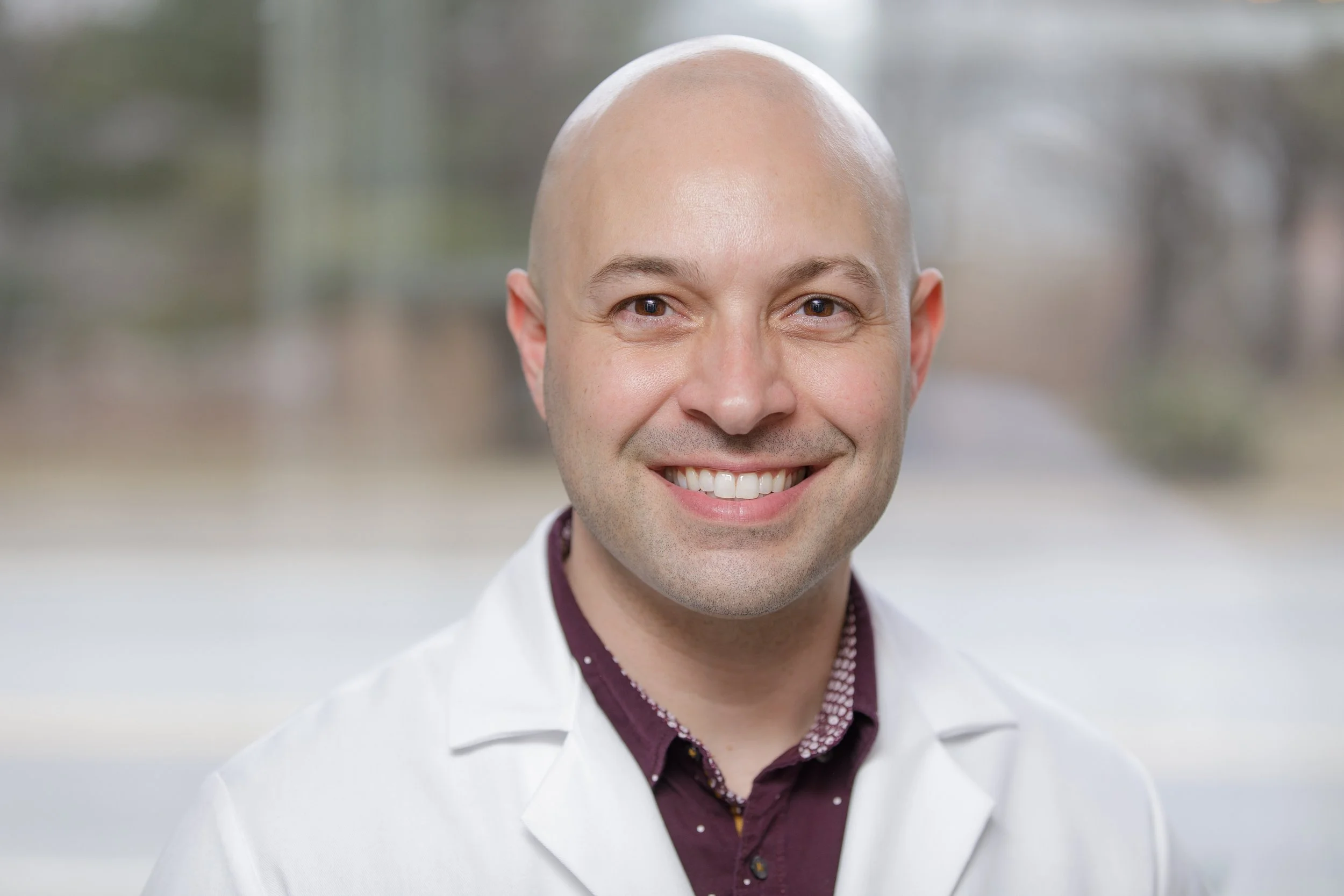 A smiling man with a shaved head, dressed in a white lab coat, standing outdoors with a blurred natural background.