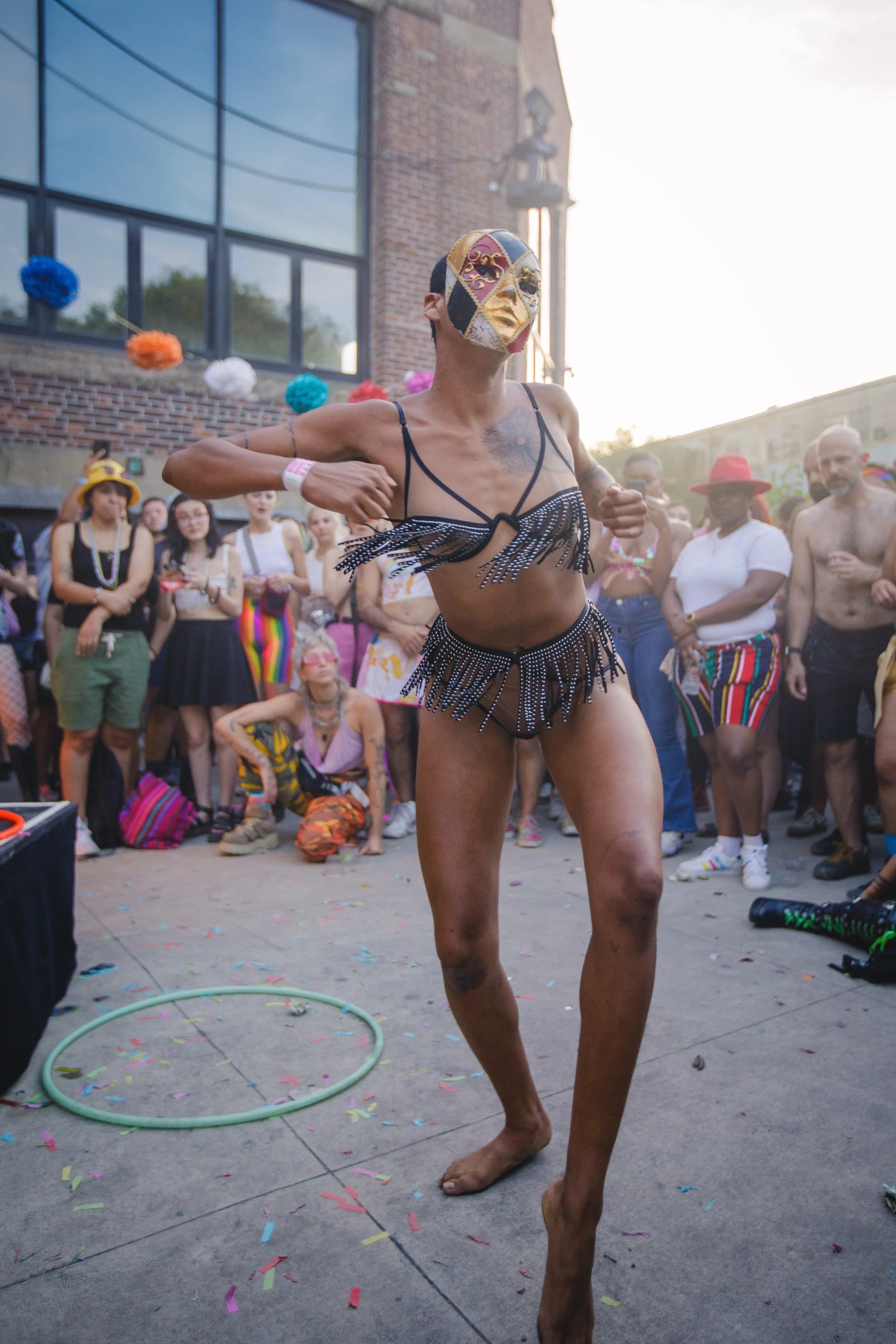 A person wearing a multi-colored mask, a black sparkly outfit with fringe, and jewelry is dancing barefoot at an outdoor event, surrounded by a crowd of spectators and colorful decorations, with confetti on the ground.