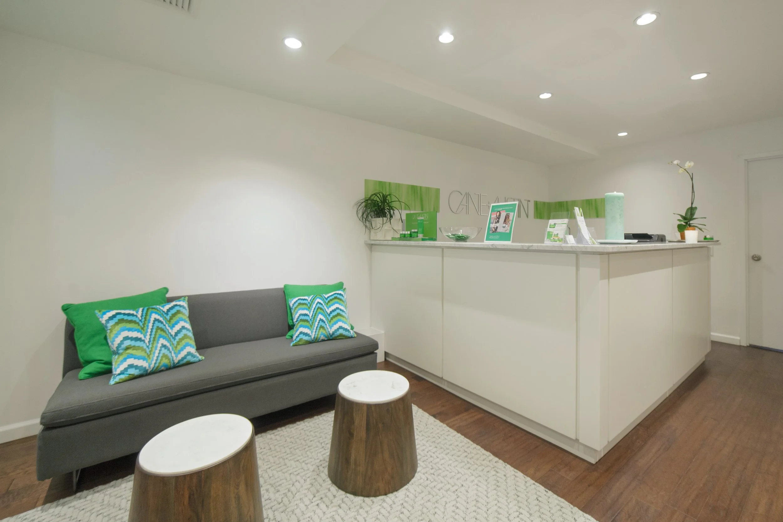 A waiting area with a gray sofa decorated with three patterned green and blue pillows, a small white round table, and two wooden cylindrical stools. The space has white walls, a cream-colored rug, and a white marble counter with green accents, plants