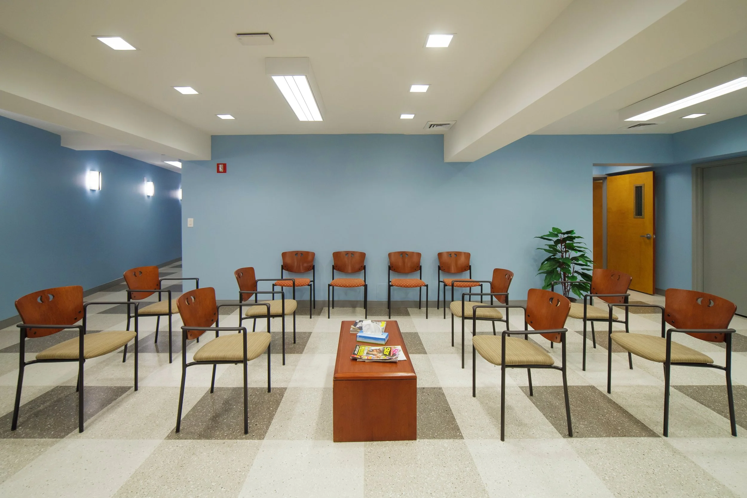 Empty waiting room with orange chairs arranged in two rows, a small wooden table with magazines, blue walls, a potted plant, and a wooden door in the background.