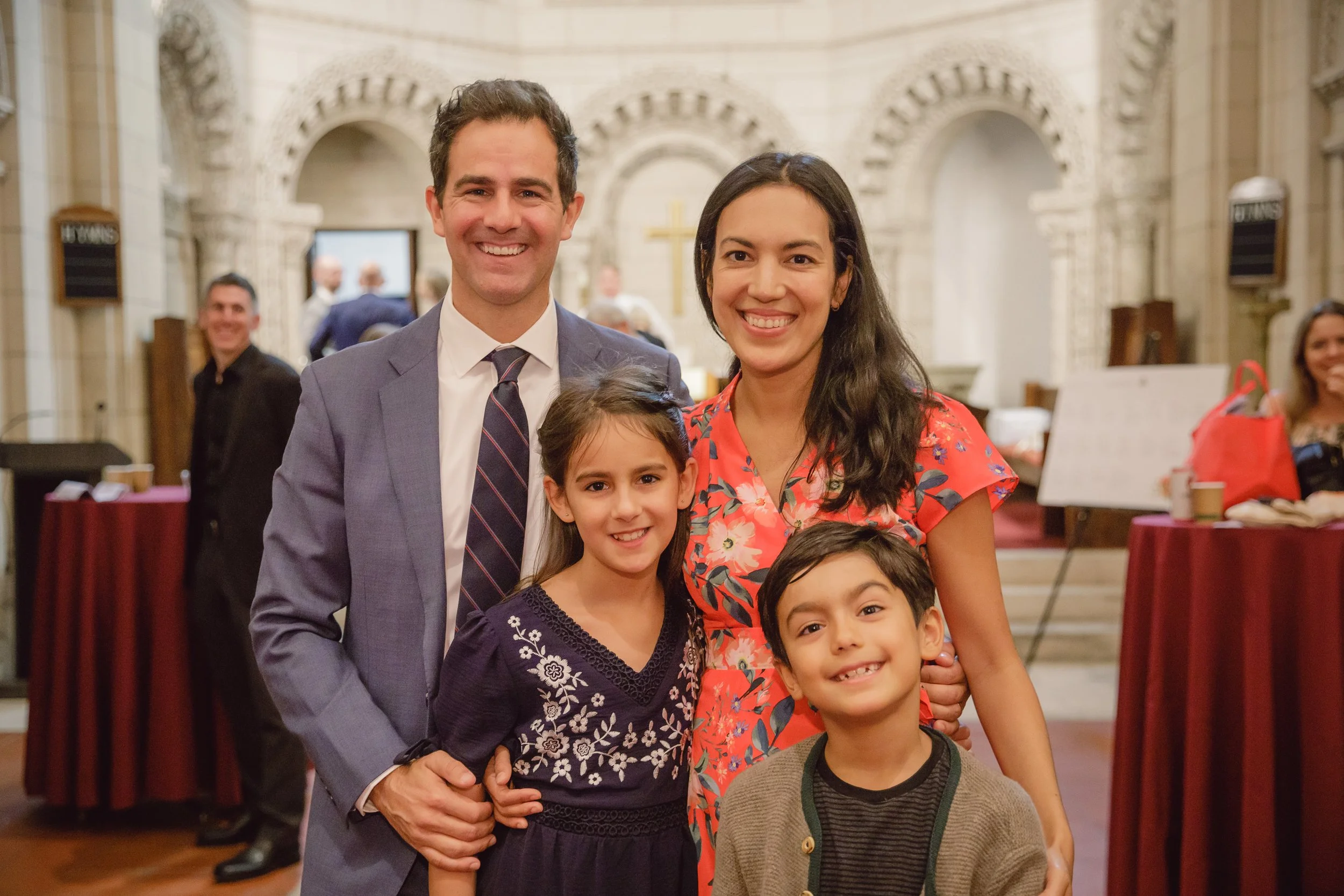 Family of five posing inside a church, with a man in a suit, a woman in a floral dress, and three children, smiling at the camera.