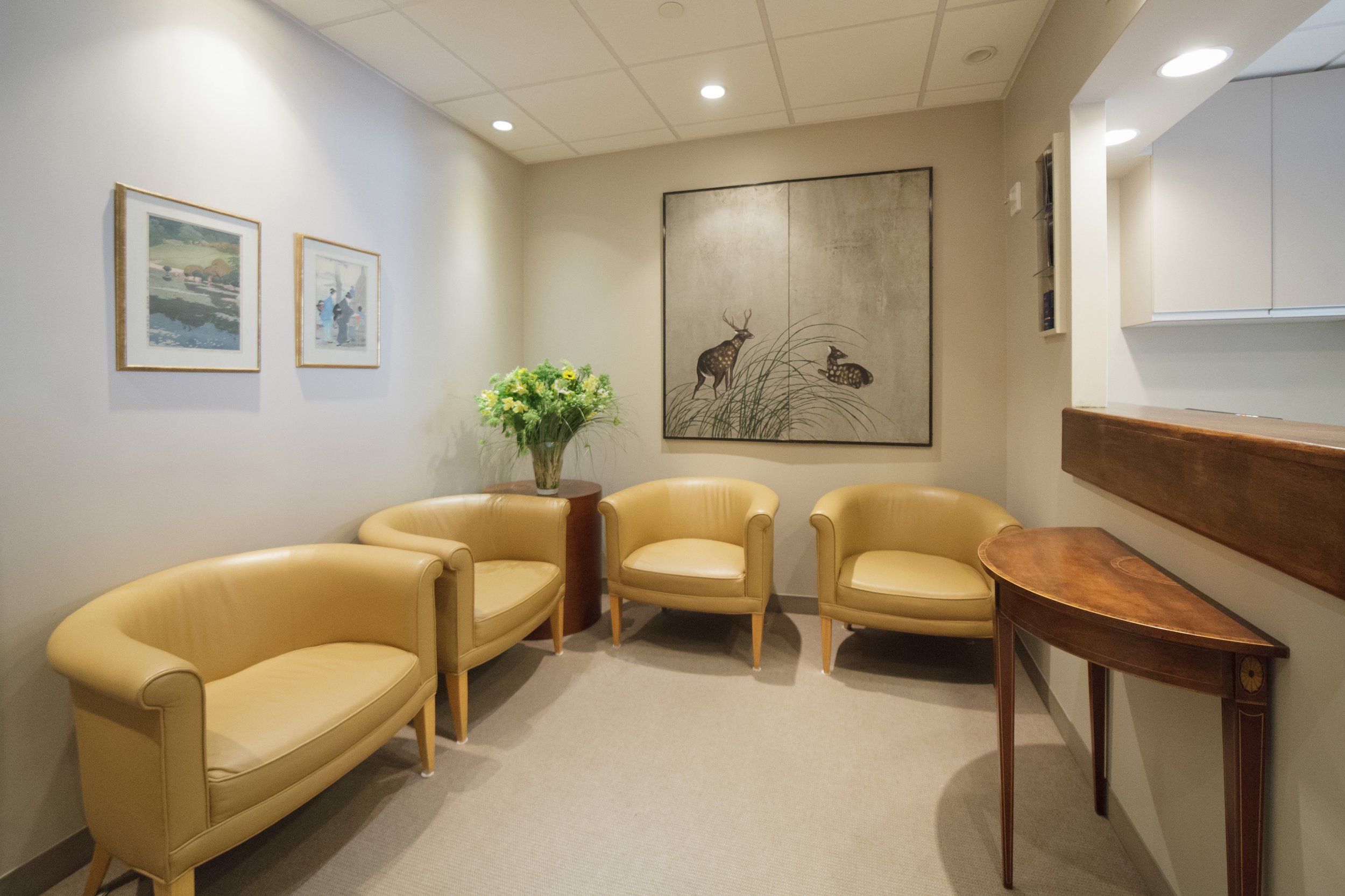 A waiting area with yellow armchairs, a round wooden table with a floral arrangement, framed artwork on the wall, and a large painting of deer and grass