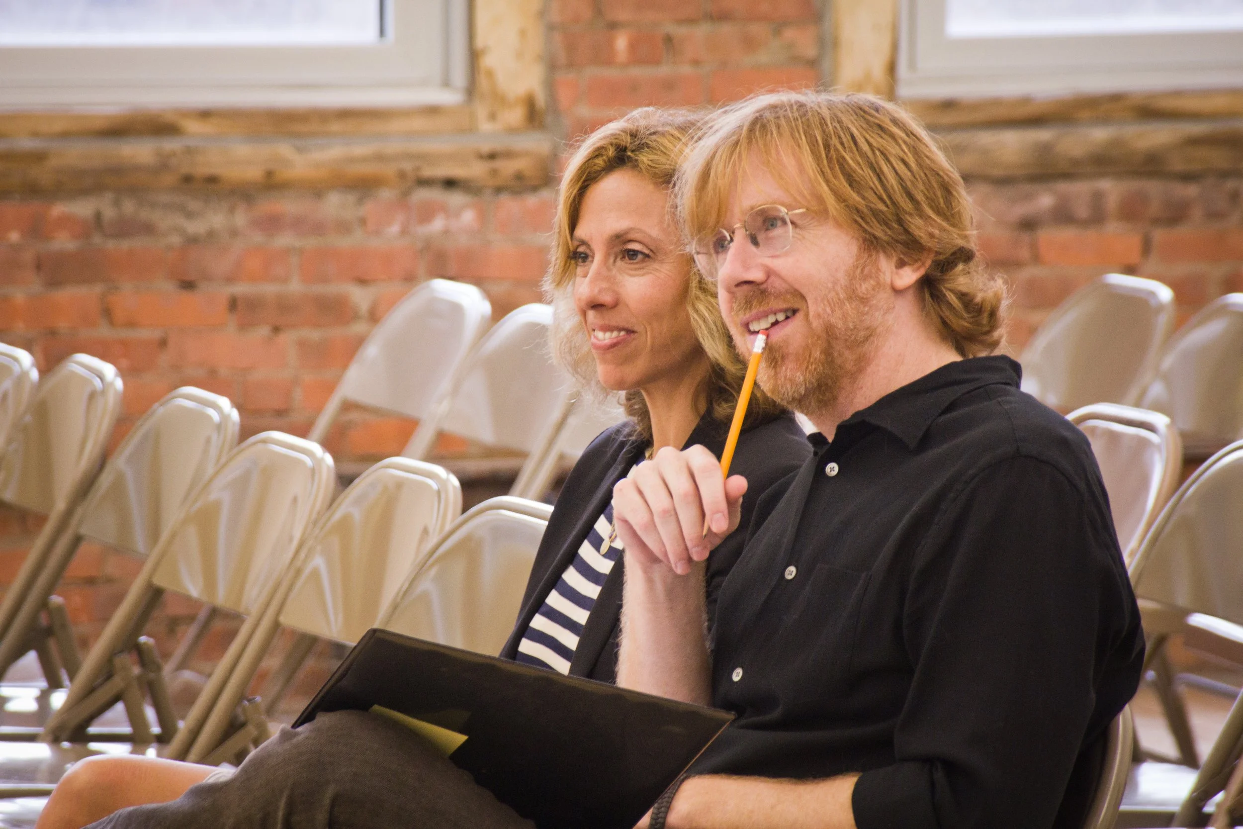 Two smiling people sitting in chairs, a woman with shoulder-length curly hair and a man with ginger hair and glasses, in a room with an exposed brick wall.