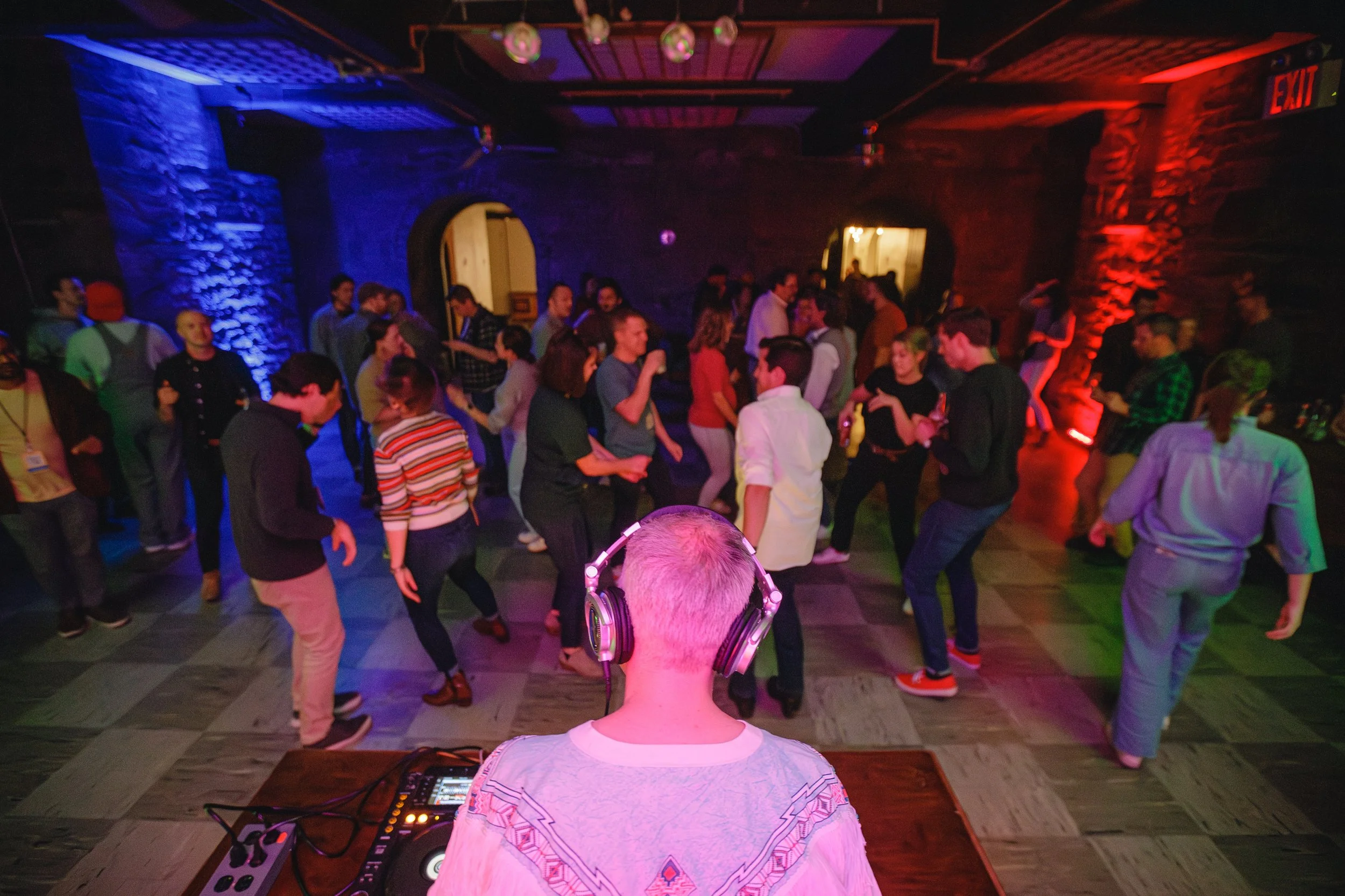 A DJ with headphones standing in front of a crowd dancing at a party or nightclub with colorful blue, red, and purple lighting.