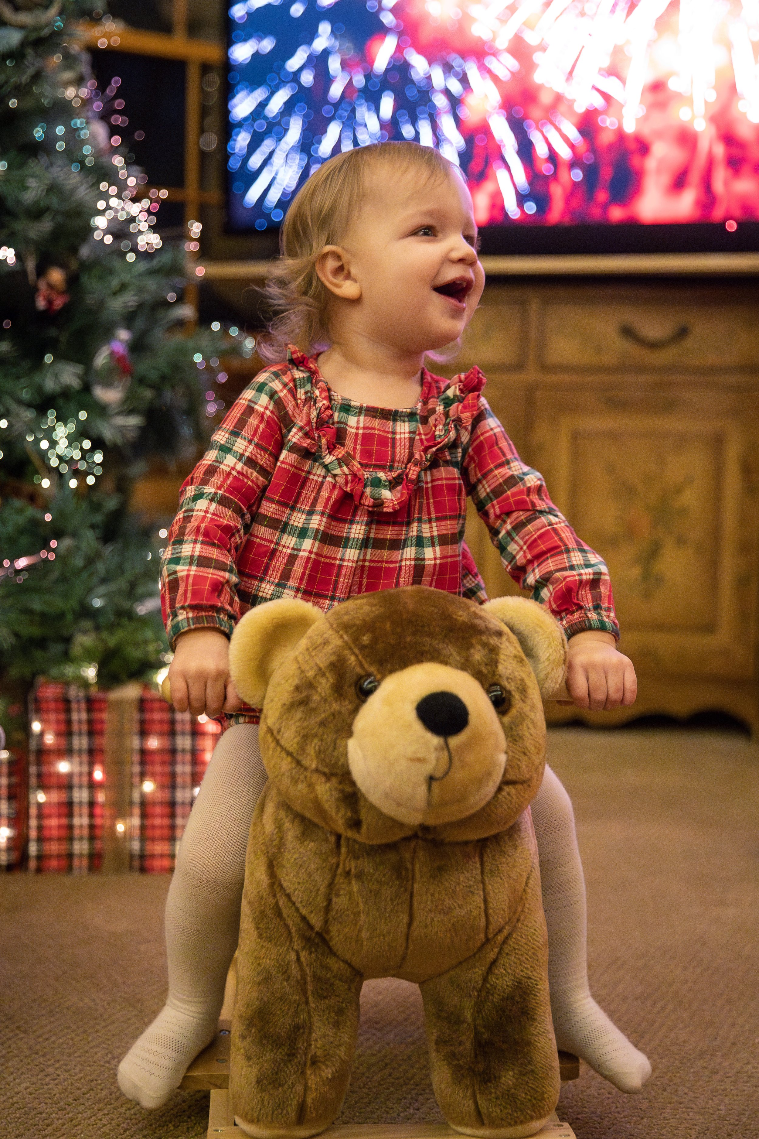 A young girl in a red plaid dress with ruffled neckline, sitting on a teddy bear ride, smiling. In the background are Christmas decorations, a Christmas tree with lights, and a TV screen displaying fireworks.