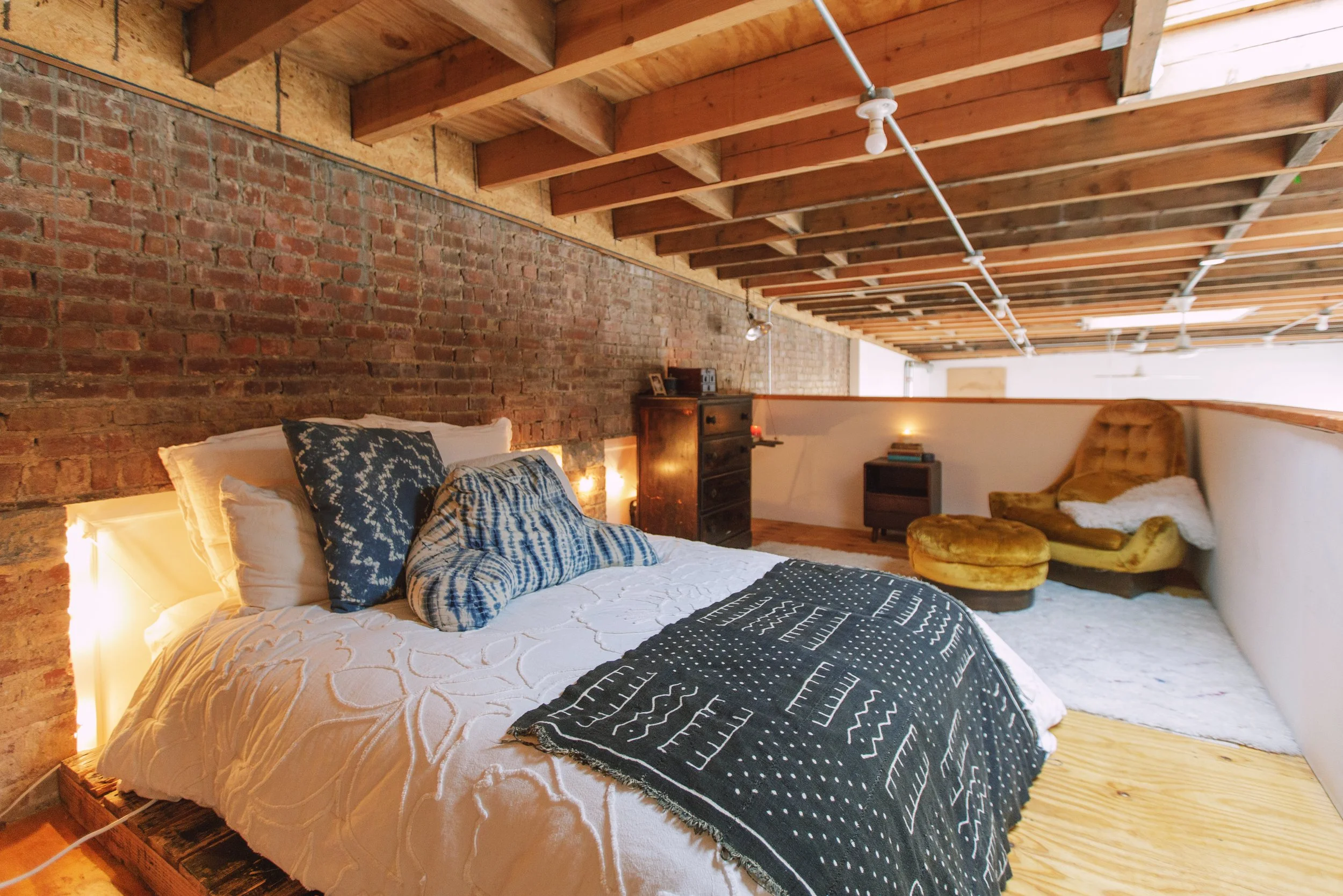 A cozy bedroom with a brick wall and exposed wooden ceiling beams. The bed has white bedding, patterned pillows, and a black and white striped throw blanket. There is a wooden dresser and a small chair or bench with a fur throw on it. The room has wa
