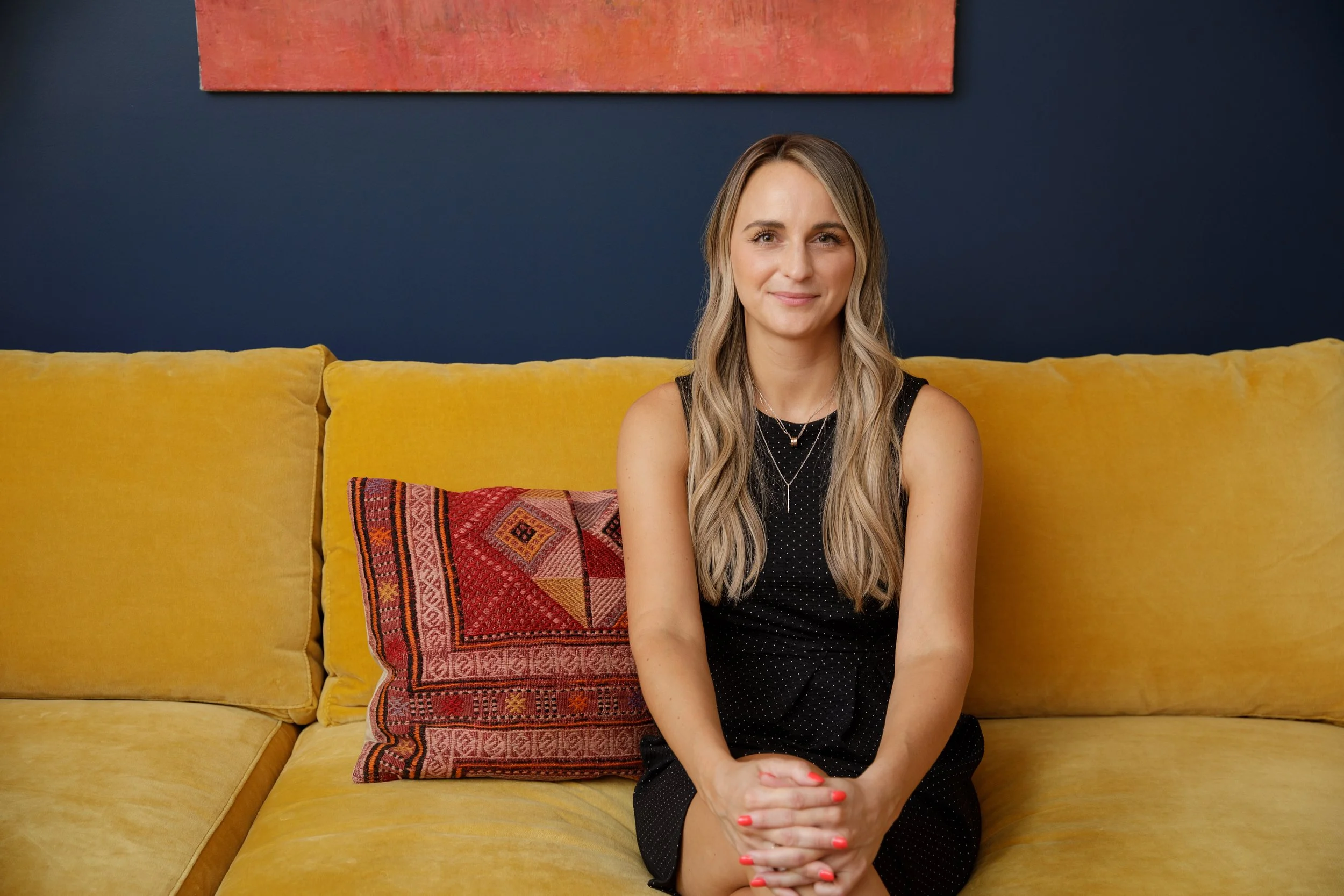 A woman with long wavy blonde hair sitting on a yellow couch with a colorful textile pillow, against a navy blue wall with a red abstract painting.