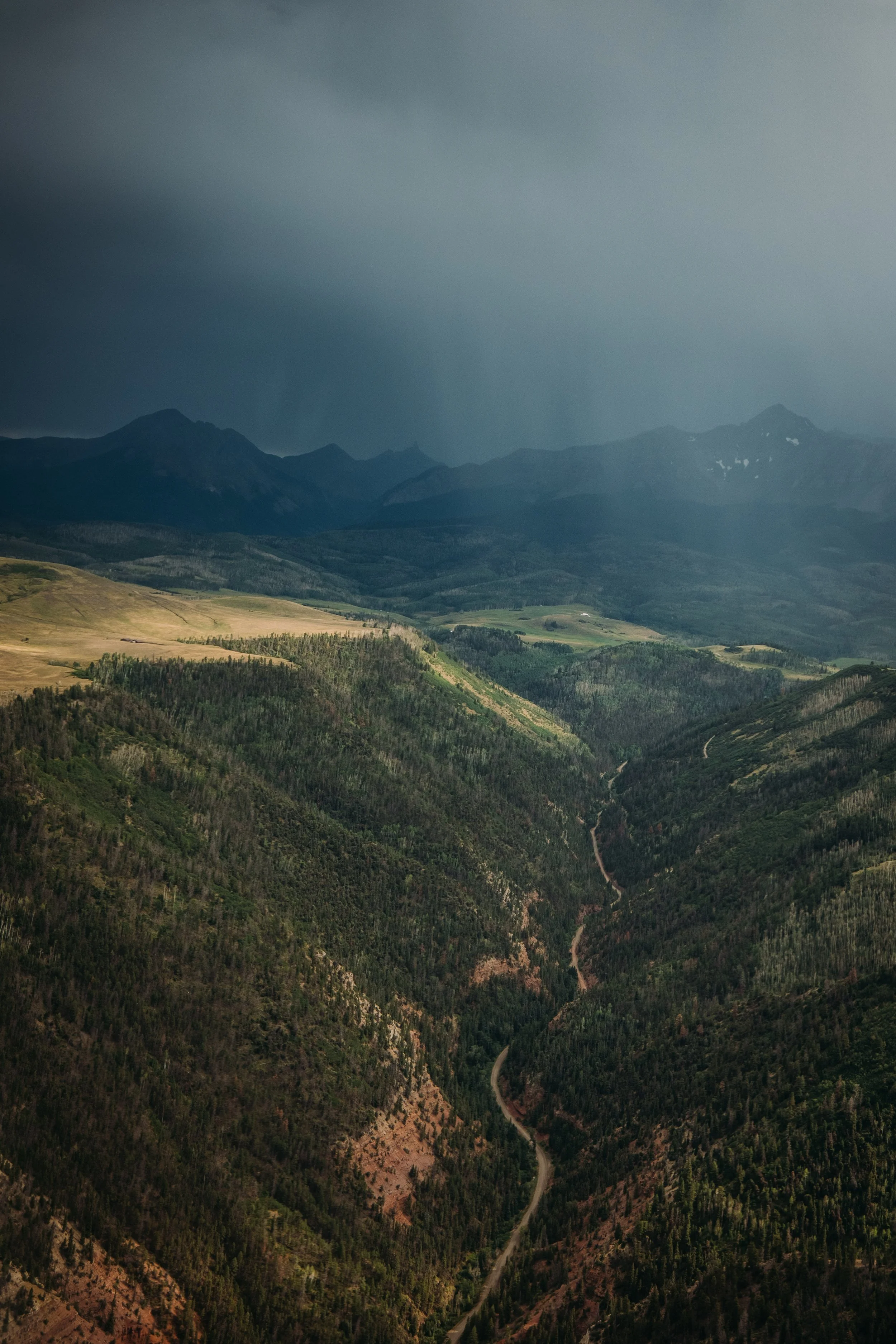 A landscape view of a mountain range with dark clouds overhead, rolling green hills, and a winding dirt road through the forest.