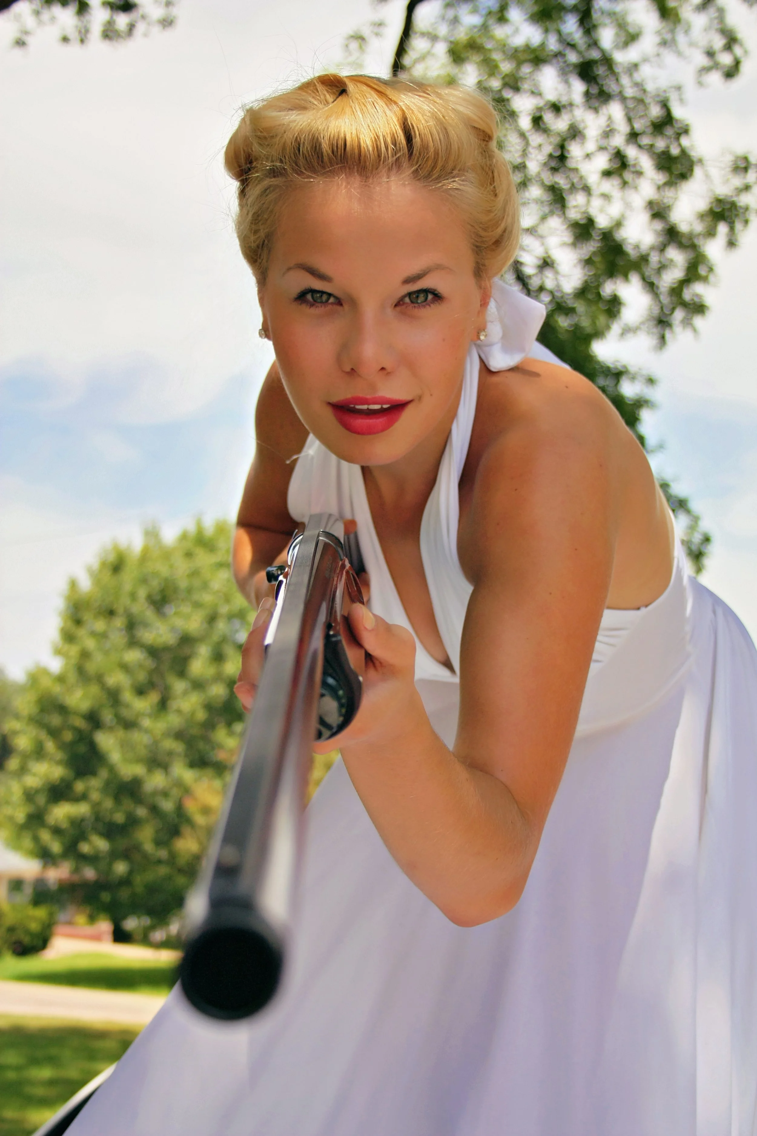 Close-up of a woman aiming a handgun outdoors with green trees and a sky in the background.