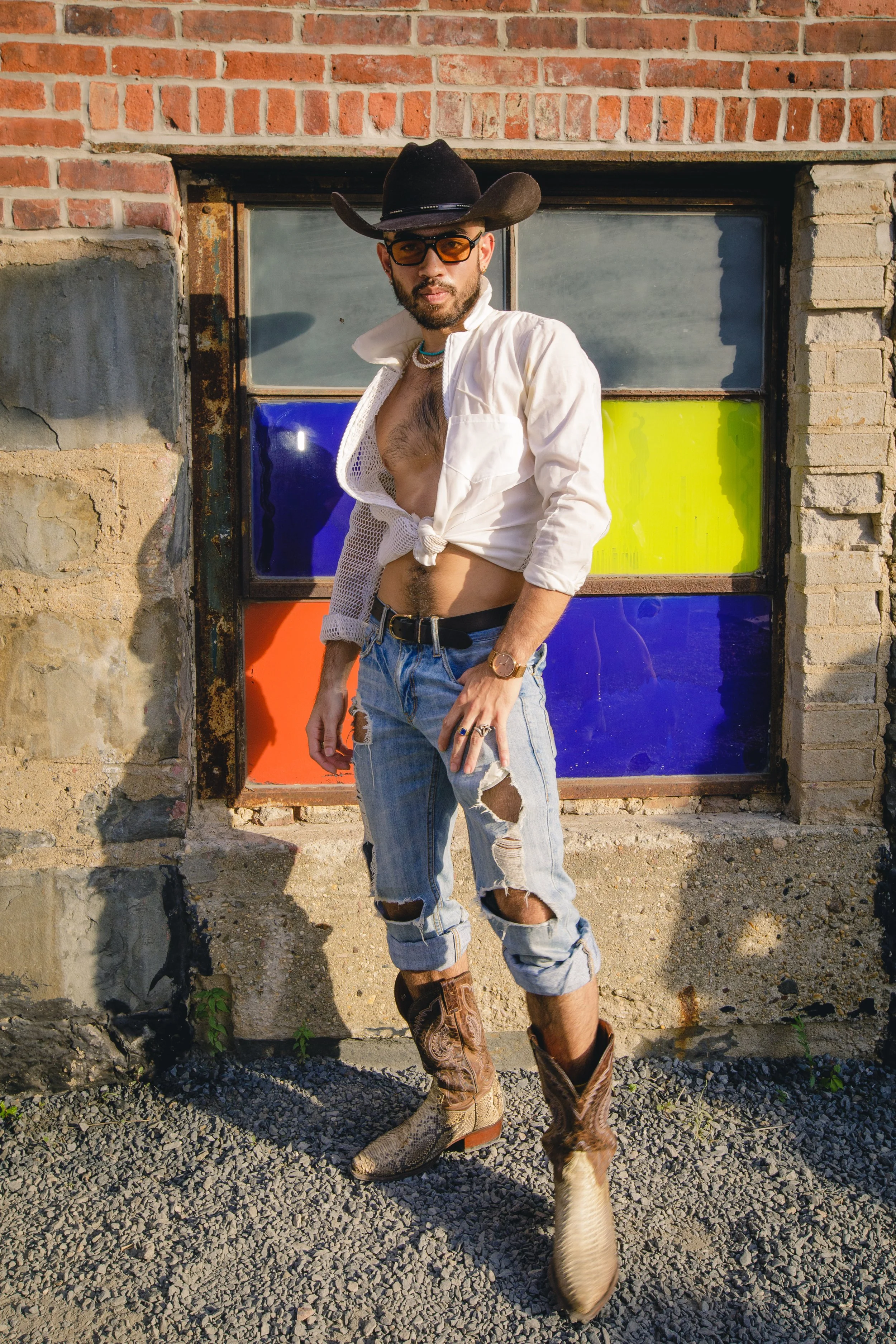 A man wearing a cowboy hat, sunglasses, and a partially unbuttoned white shirt revealing chest hair, standing outdoors in front of a building with colorful window panes. He has ripped jeans and cowboy boots, and there is a dog with a light-colored co