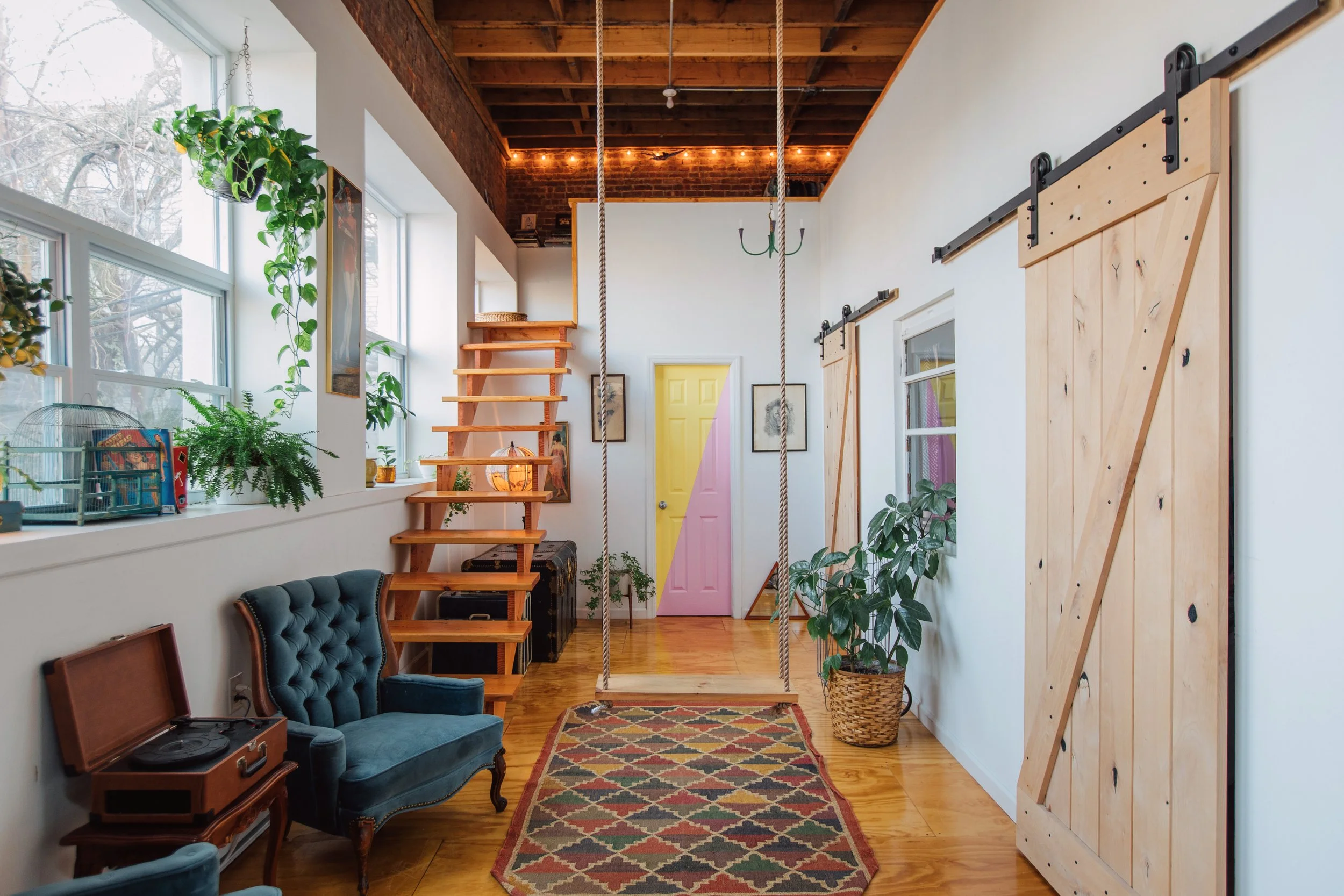 Interior of a cozy living space featuring large windows, houseplants, a vintage record player, velvet armchair, a colorful rug, a yellow and pink door, wooden stairs, a climbing rope, and decorative lighting with string lights.