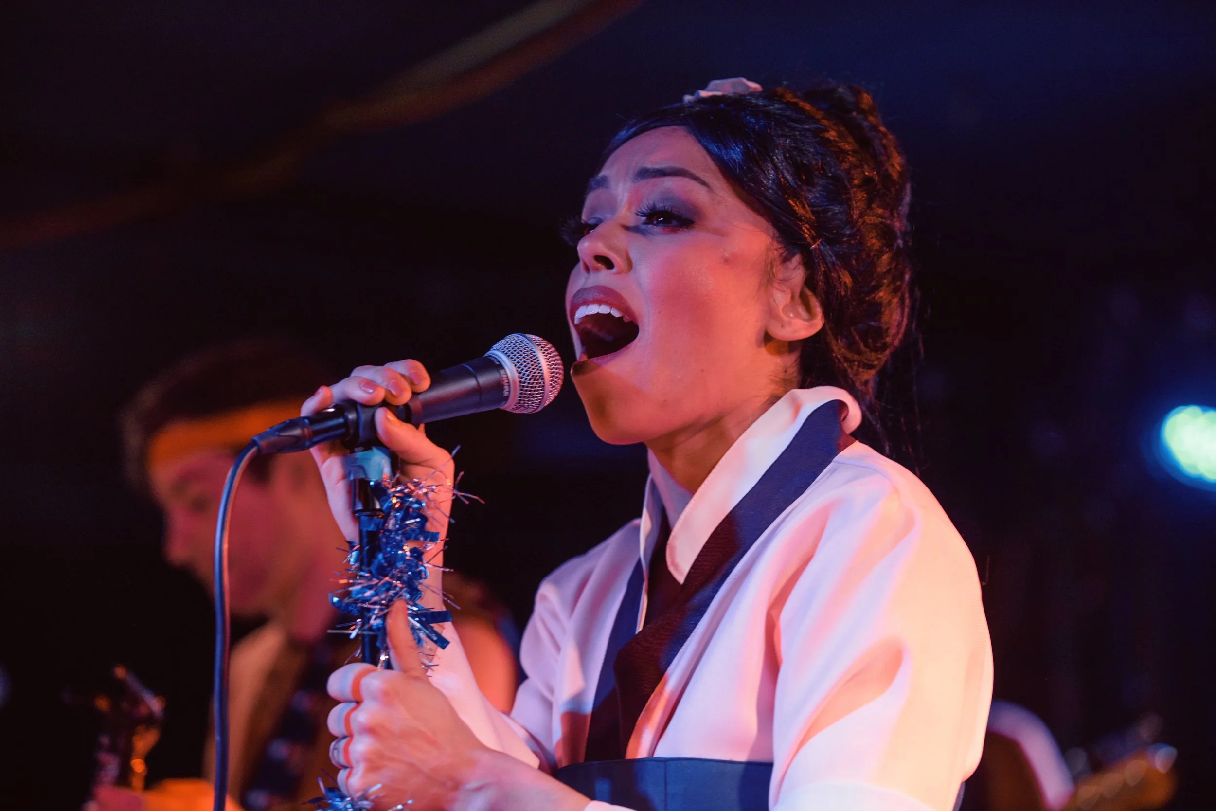A woman singing into a microphone during a performance, wearing a white outfit with dark accents, with colorful stage lighting in the background.