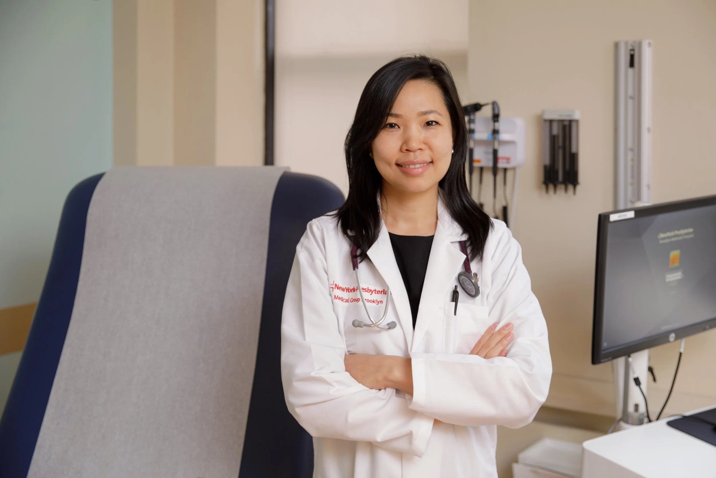 A smiling female doctor in a white coat and stethoscope in a medical office, with her arms crossed, looking at the camera.