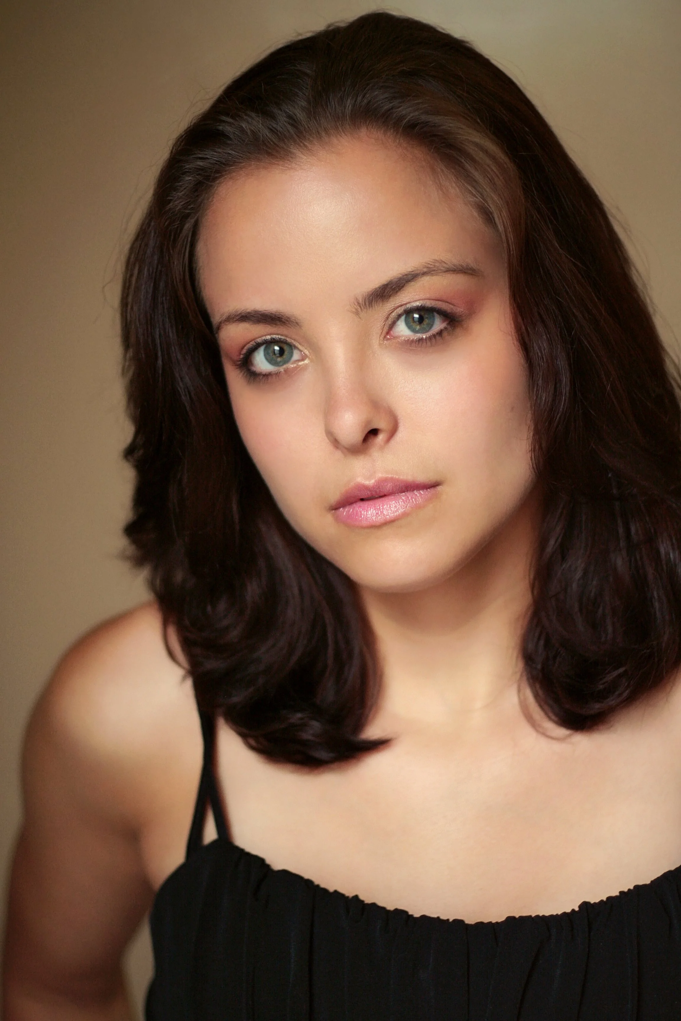 A woman with long, dark wavy hair and light skin, wearing a black spaghetti strap top, looking directly at the camera with neutral expression.