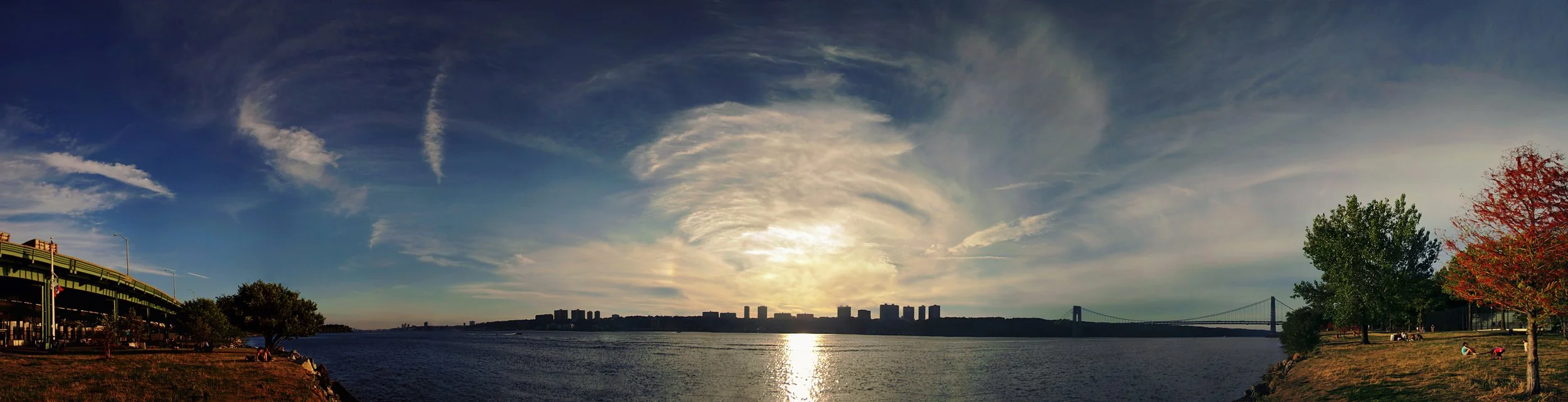 Panoramic view of a river with a city skyline, bridge, and trees during sunset or sunrise, with a partly cloudy sky.