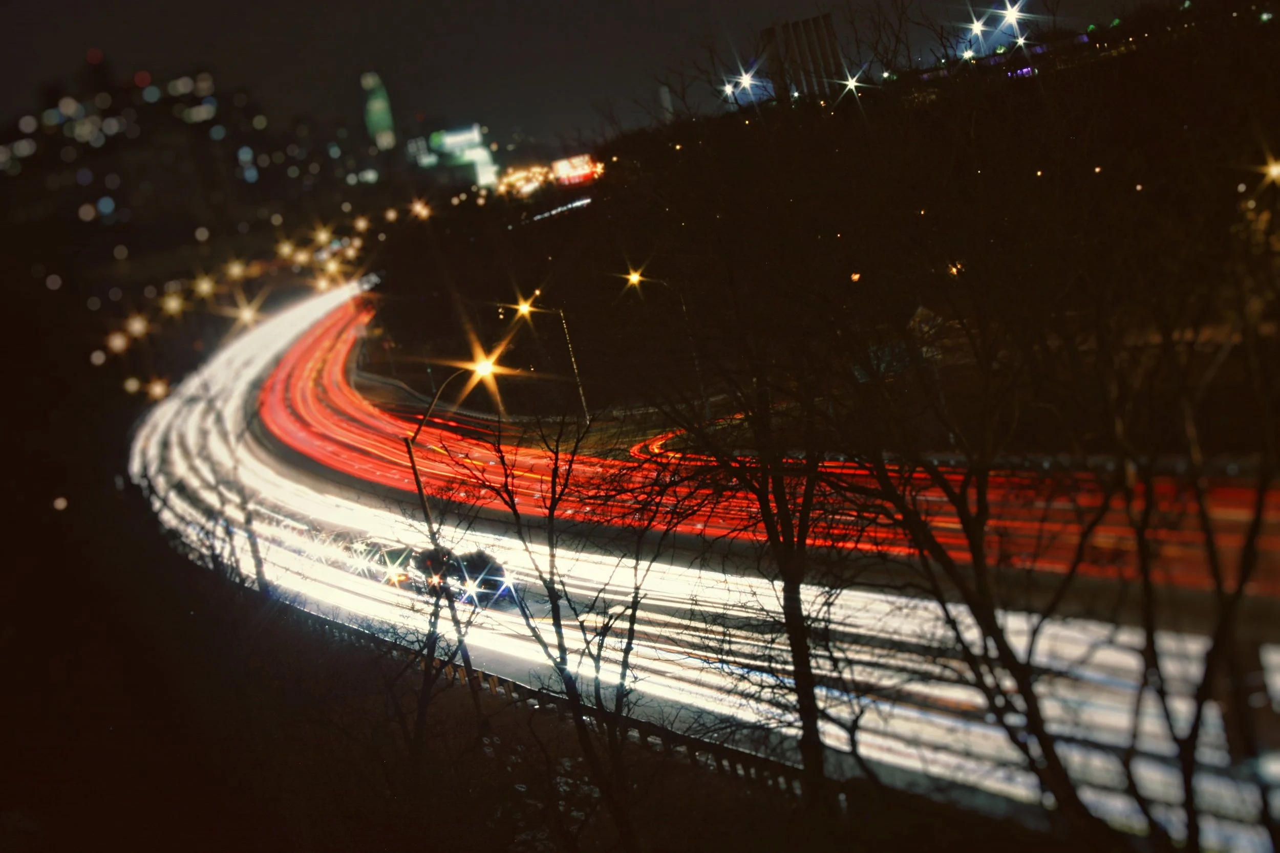 Long exposure photo of a highway at night with streaks of red and white lights from moving vehicles, city skyline in the background, and trees silhouetted in the foreground.