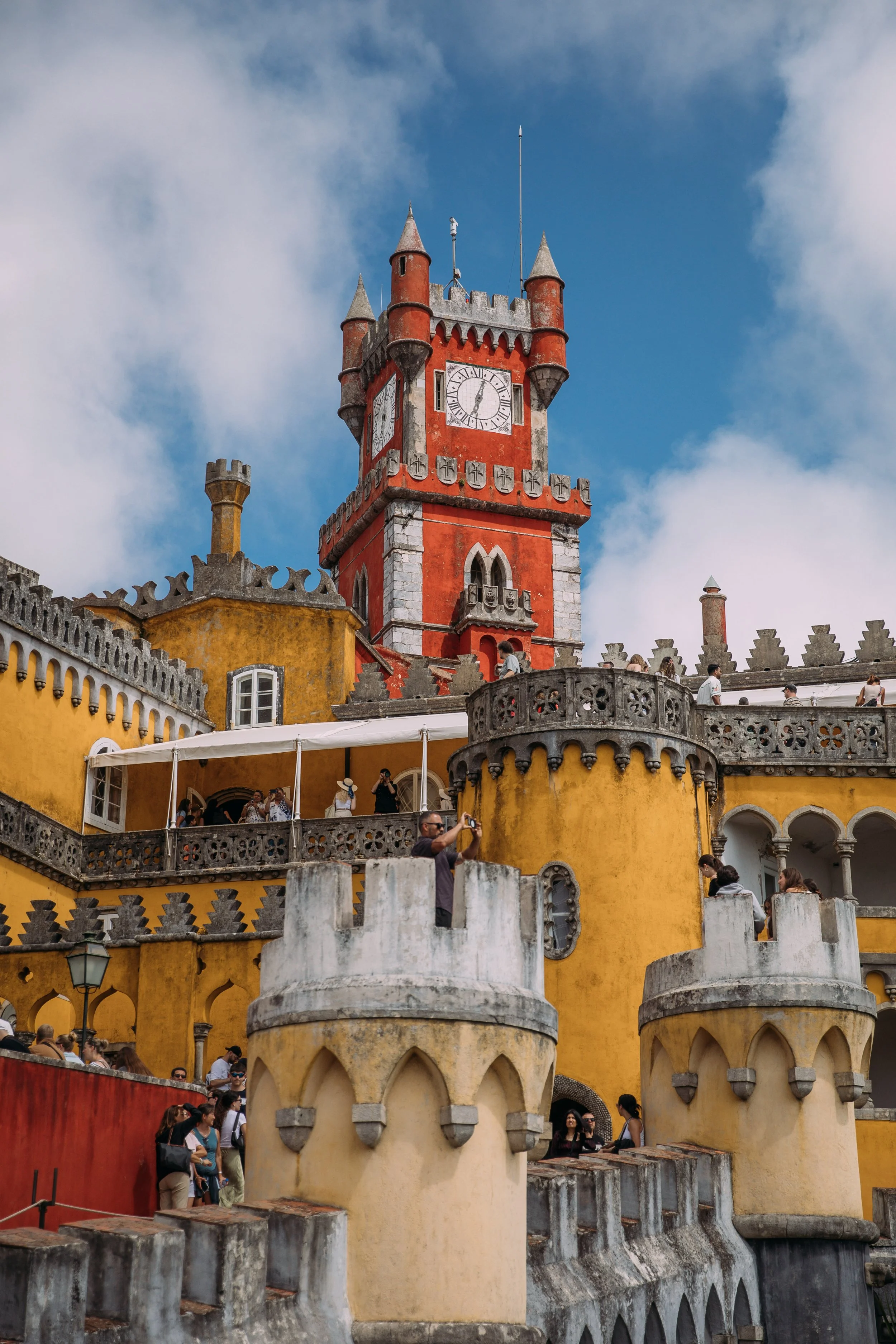 Colorful castle with a red and yellow facade, a clock tower with four small turrets, and a crowd of tourists on an overcast day.