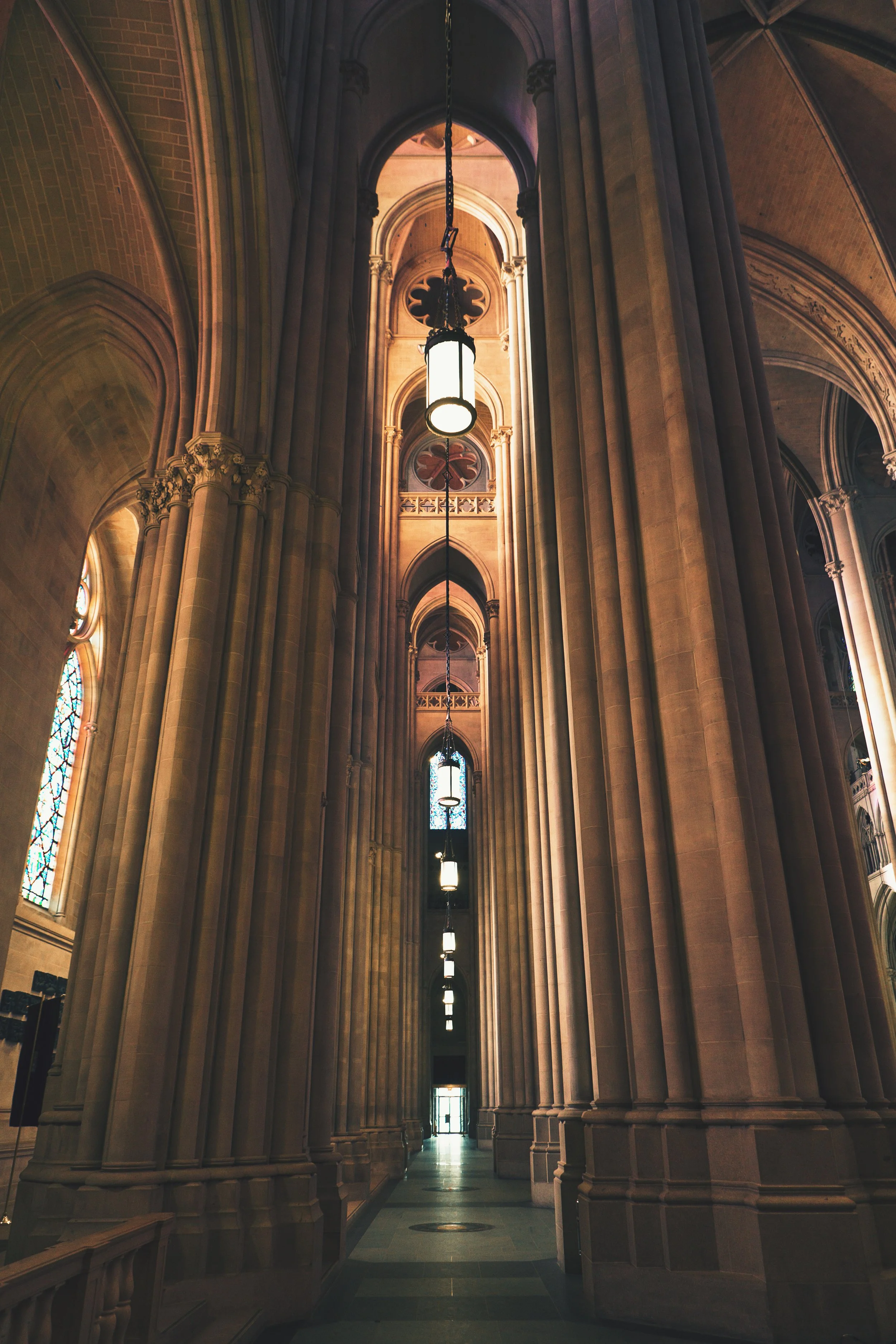 Interior of a grand cathedral with tall, ornate pillars supporting high vaulted ceilings, illuminated by hanging pendant lights, with stained glass windows along the side walls.