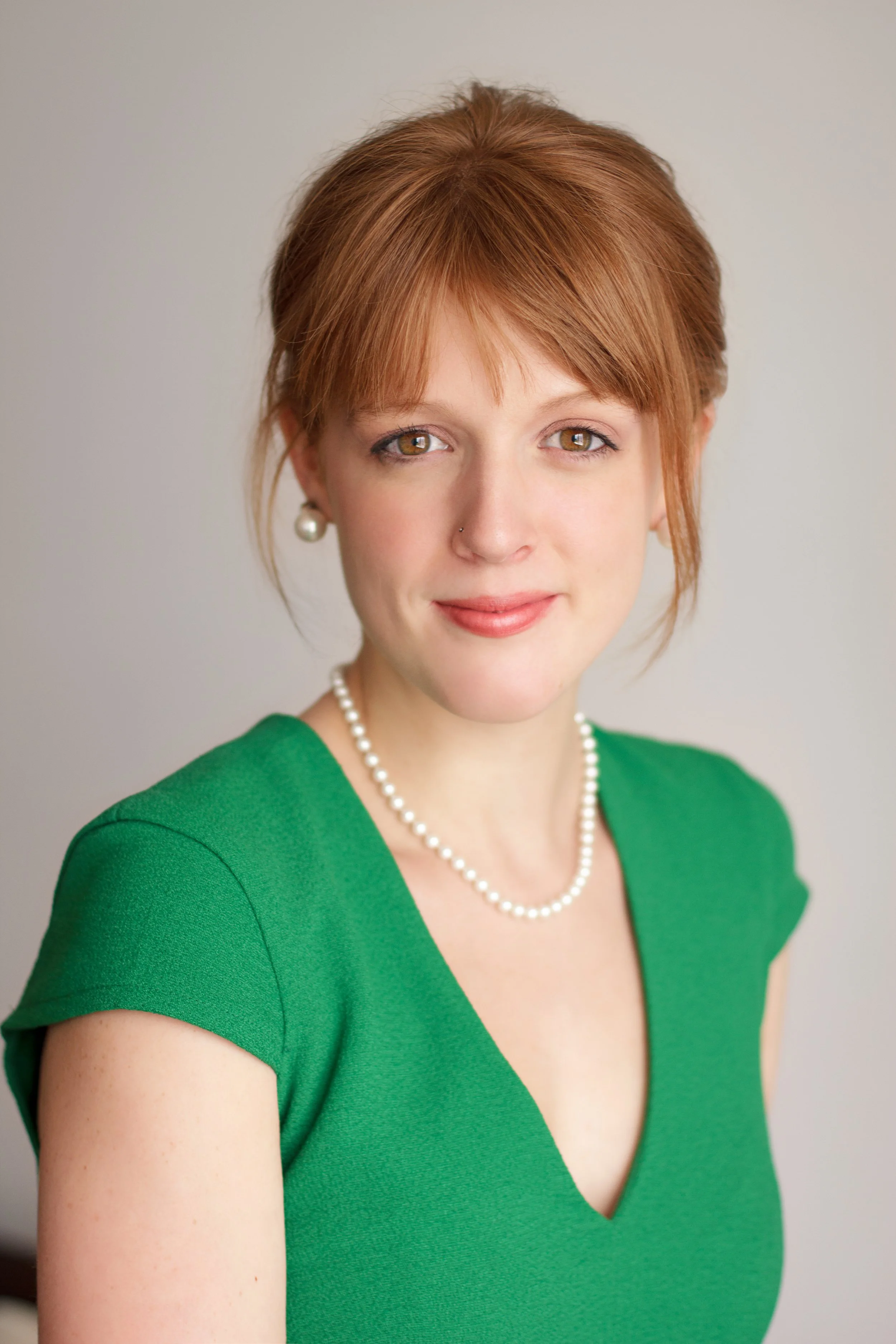 A woman with red hair styled in a loose, layered cut, wearing a green dress, pearl earrings, and a pearl necklace, smiling softly against a plain light background.