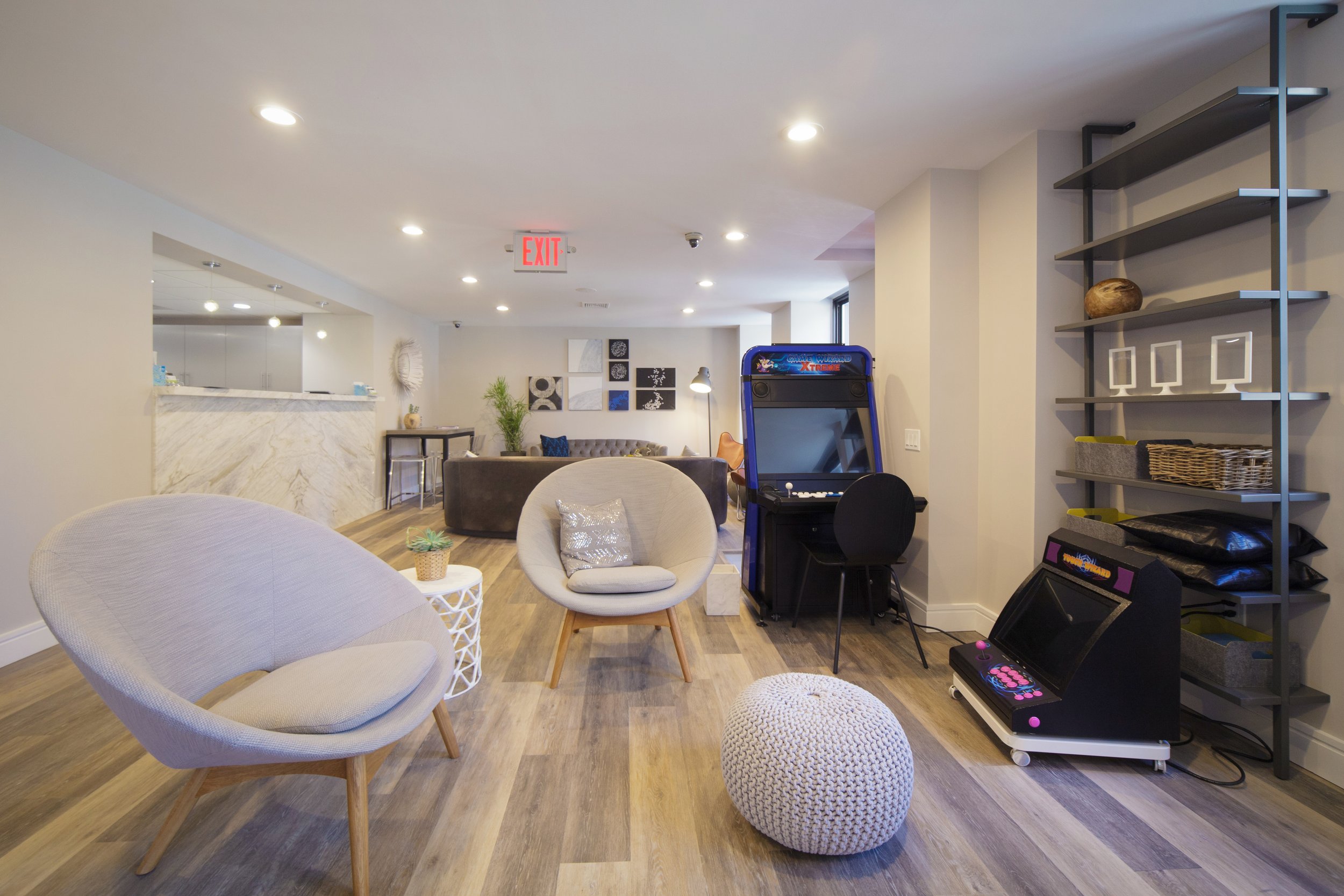 Interior view of a modern lounge area with white chairs, a small white side table, a colorful wall art, a gaming arcade machine, and a shelving unit with decor. Ceiling lights and wooden flooring are visible.