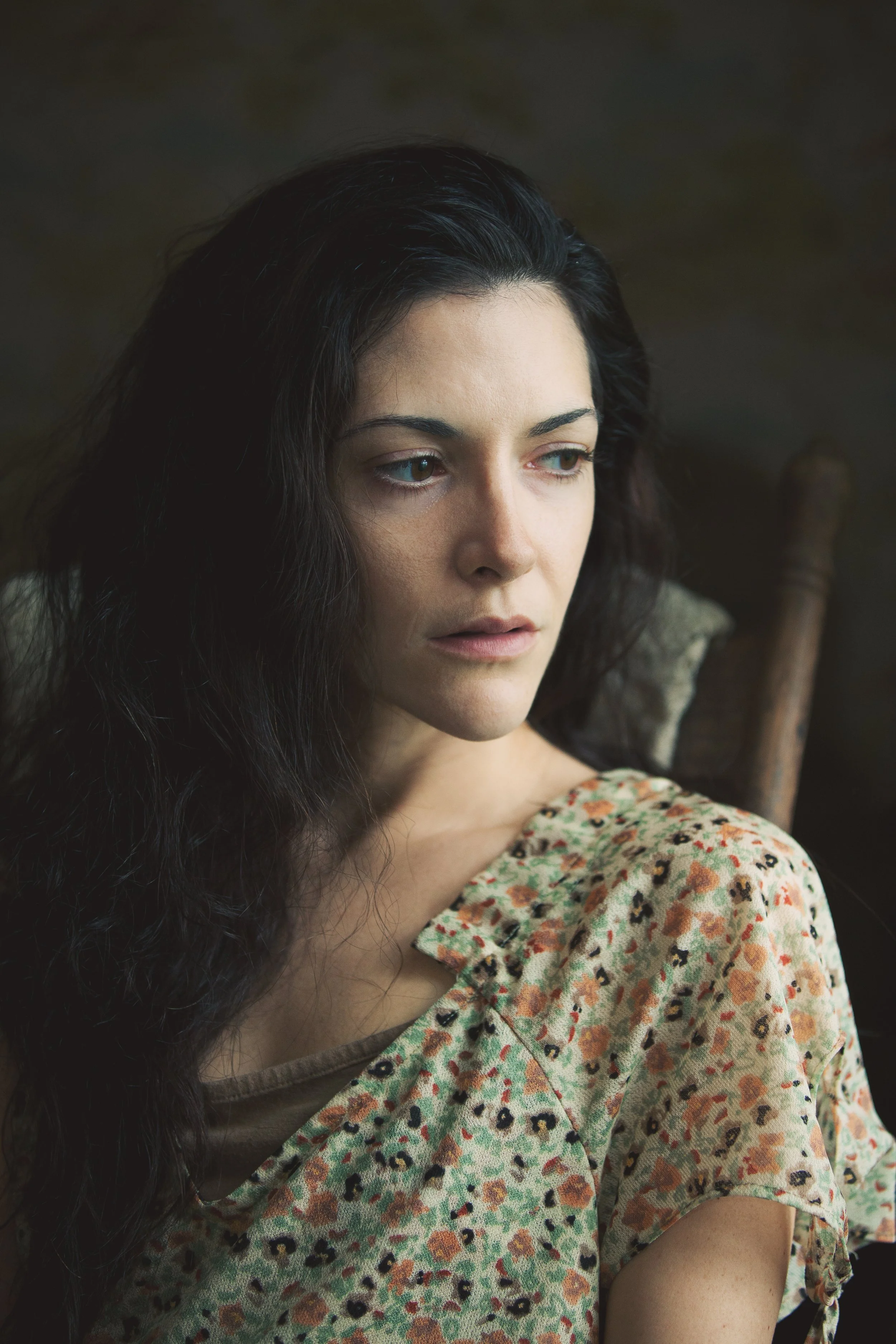 A woman with long dark hair and light skin, wearing a floral-patterned dress, looking down thoughtfully indoors.