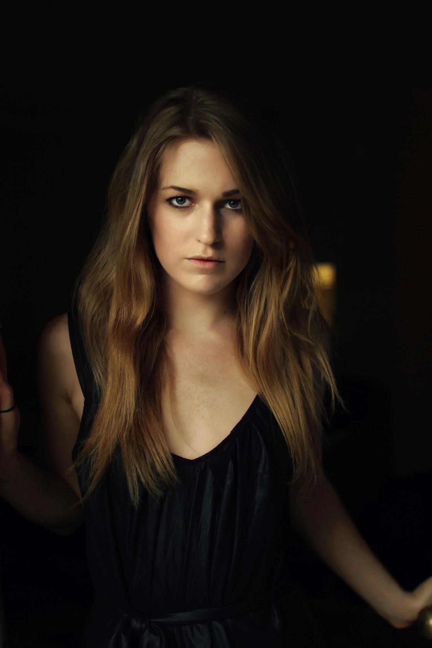A young woman with long, wavy auburn hair and blue eyes, wearing a black sleeveless top, stands against a dark background with a subtle light source illuminating her face.