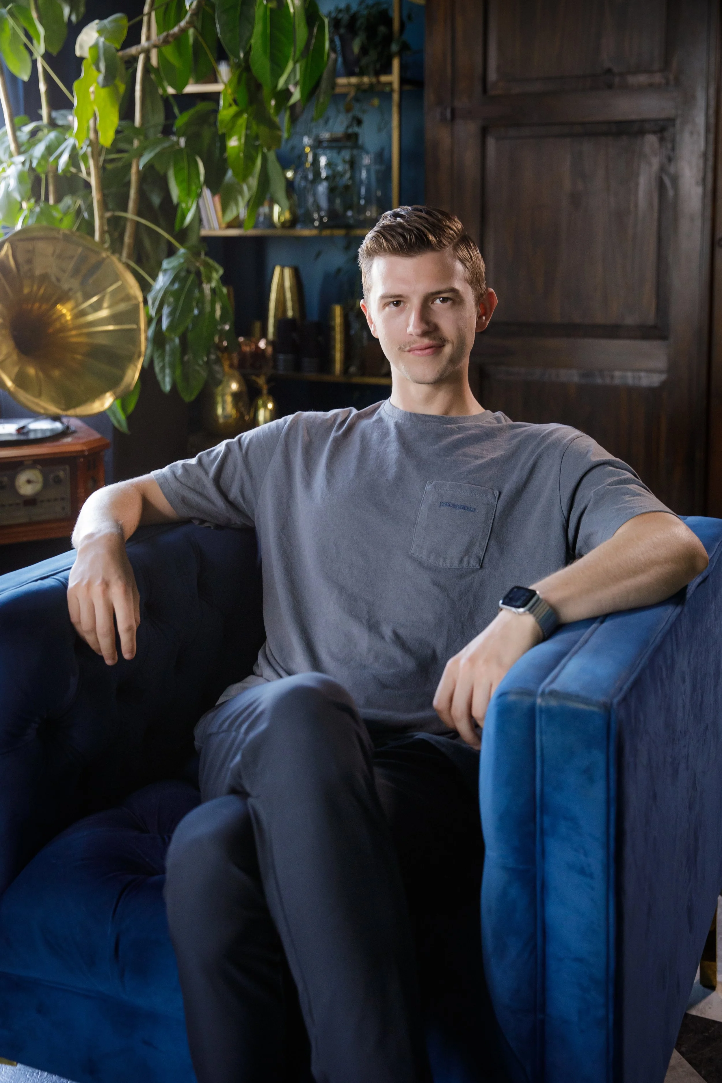 A young man sitting relaxed on a blue velvet sofa in a room with dark wooden paneling and large green potted plant. He is wearing a casual gray t-shirt and a smartwatch.