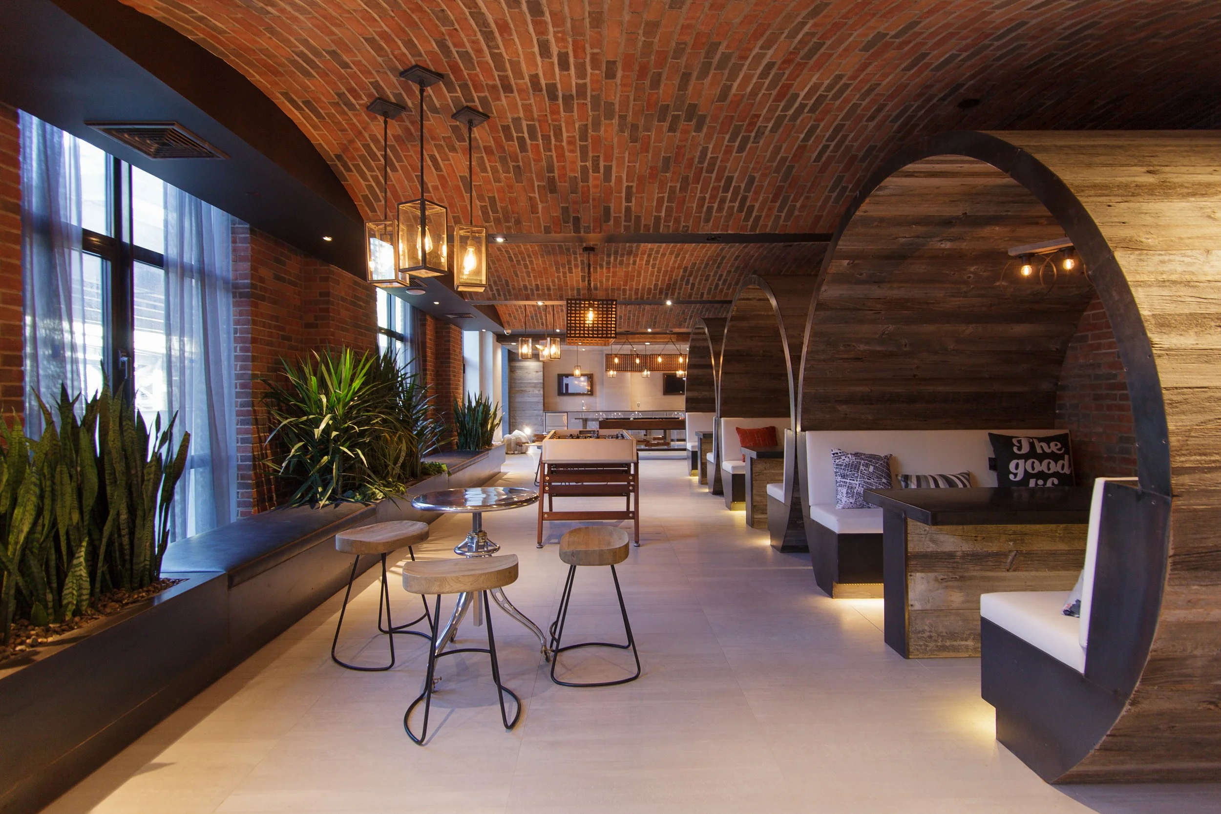 Interior of a modern restaurant or cafe with a brick vaulted ceiling, hanging light fixtures, large windows with sheer curtains, and various seating options including tables, chairs, and built-in bench seats with pillows.