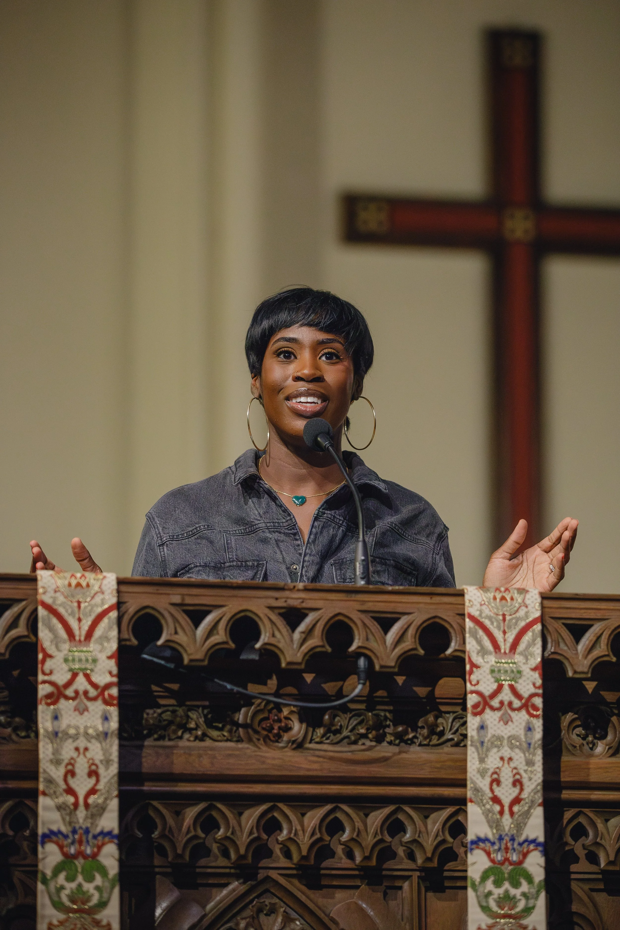 An African American woman with short black hair and hoop earrings speaking at a church pulpit with colorful banners hanging in front, a microphone, and a large cross in the background.