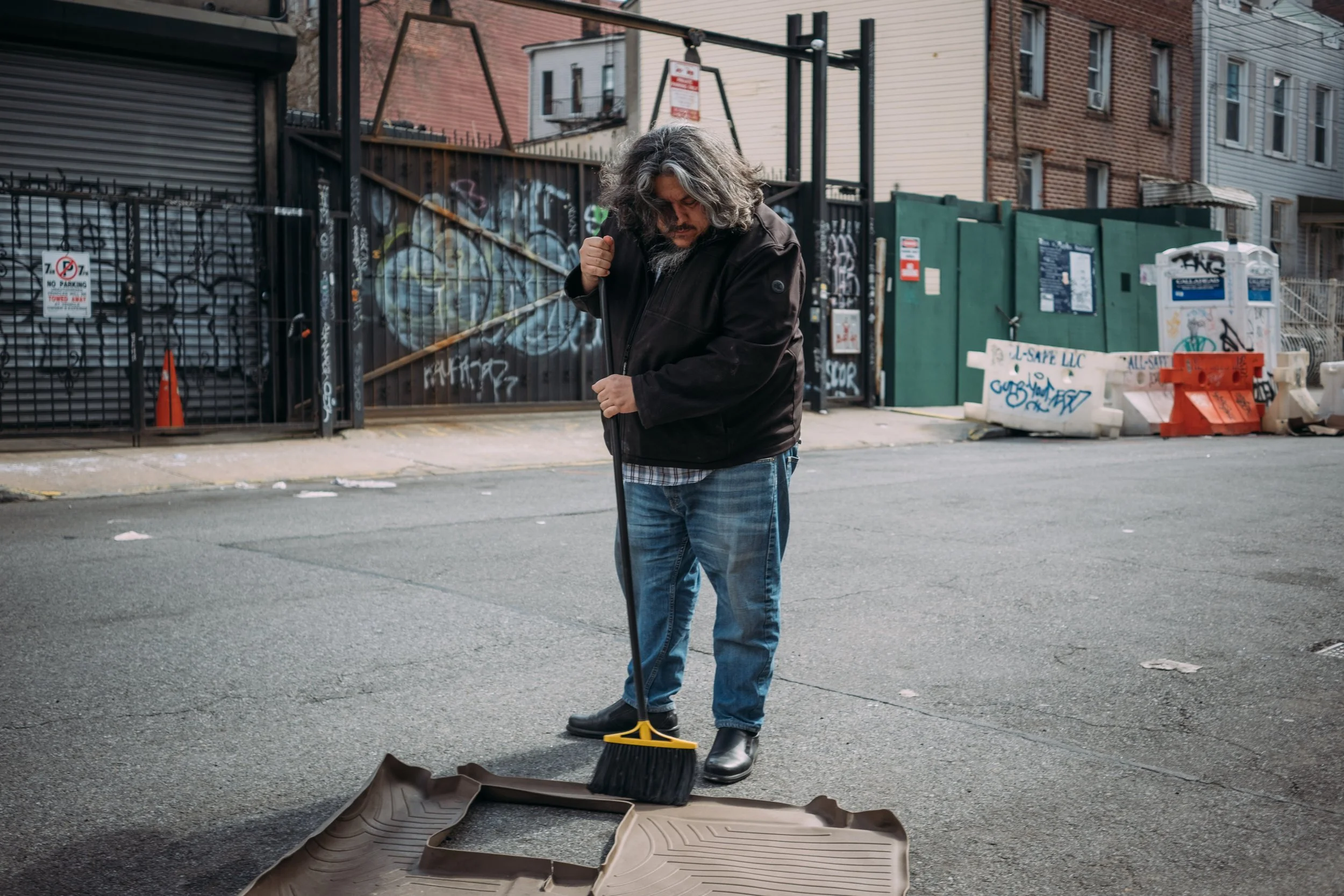 A man with long gray hair and beard sweeping debris on the street with a broom, wearing a black jacket, plaid shirt, and jeans.