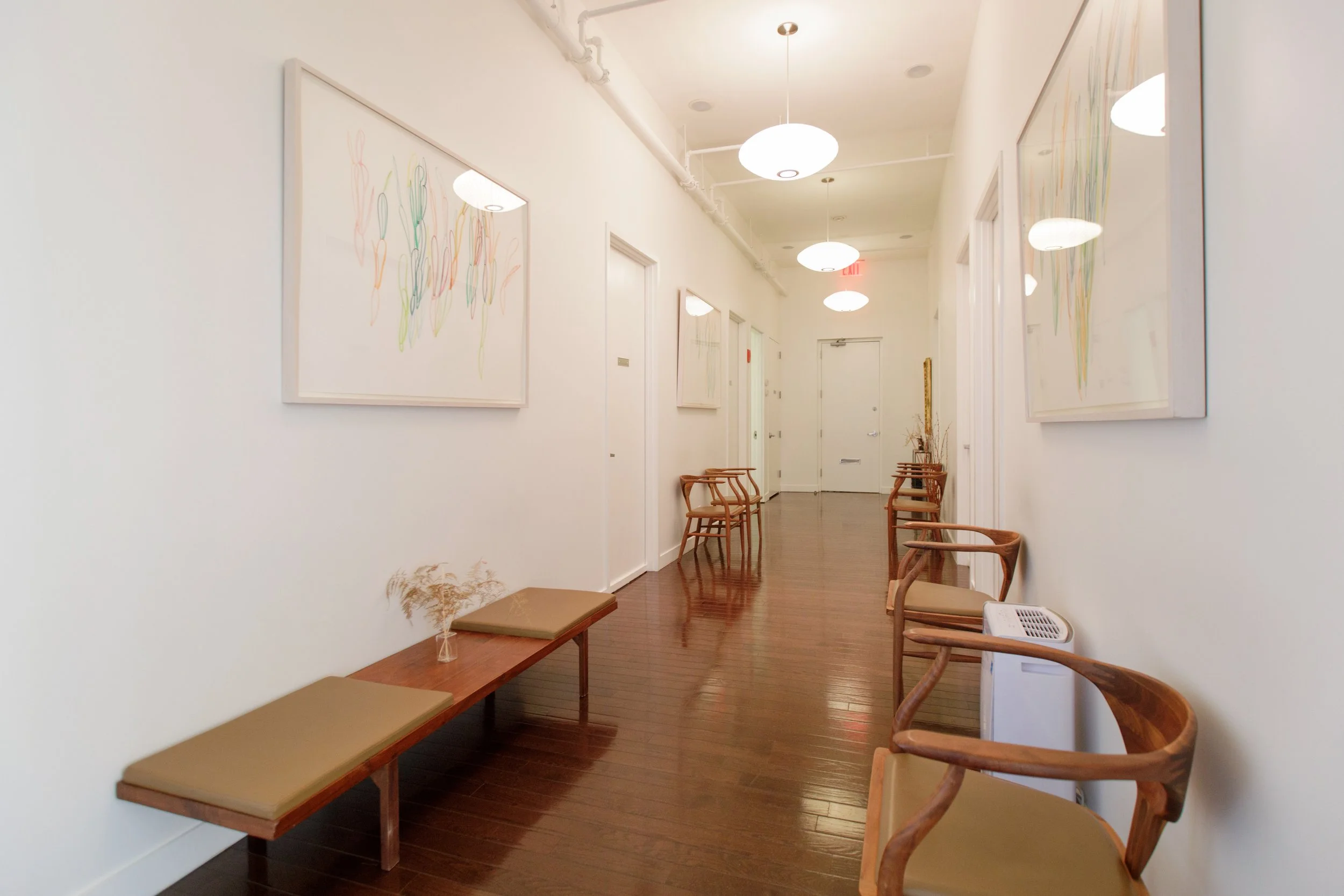 A hallway with white walls, wooden floors, and multiple framed abstract artworks. There are several wooden chairs with beige cushions along the wall and a bench with beige cushions and a small vase with dried plants on it. Ceiling lights and an exit 