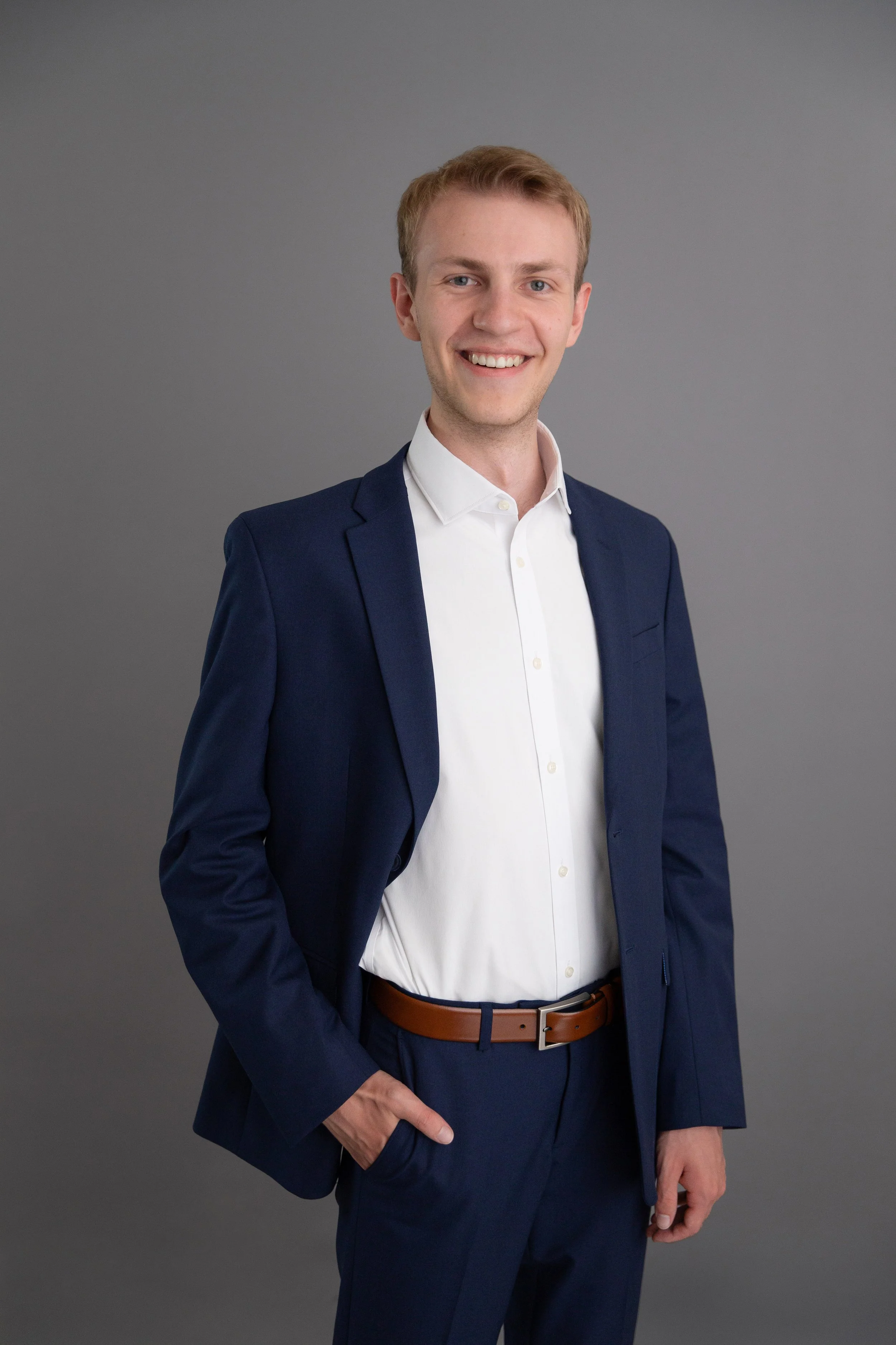 Portrait of a young man wearing a dark blue suit, white shirt, and brown belt, smiling against a plain gray background.