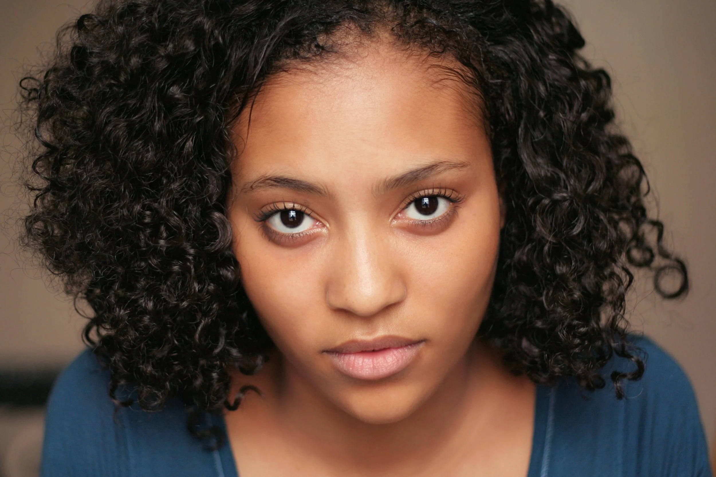 Close-up of a young woman's face with dark, curly hair and expressive eyes, looking directly at the camera.