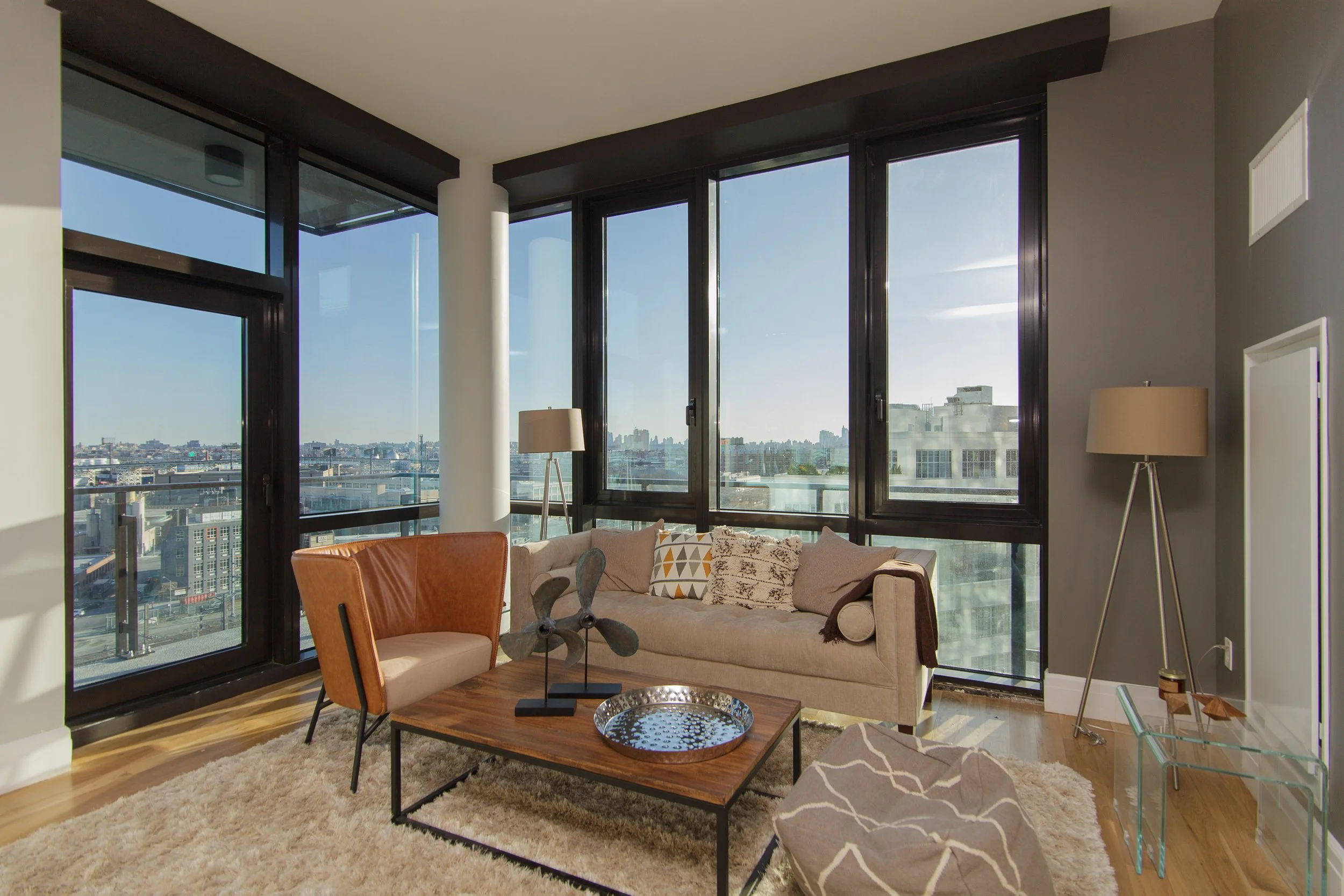 Living room with large windows showing city skyline, beige sofa with geometric pillows, orange armchair, coffee table with decorative items, floor lamp, area rug, and modern decor.