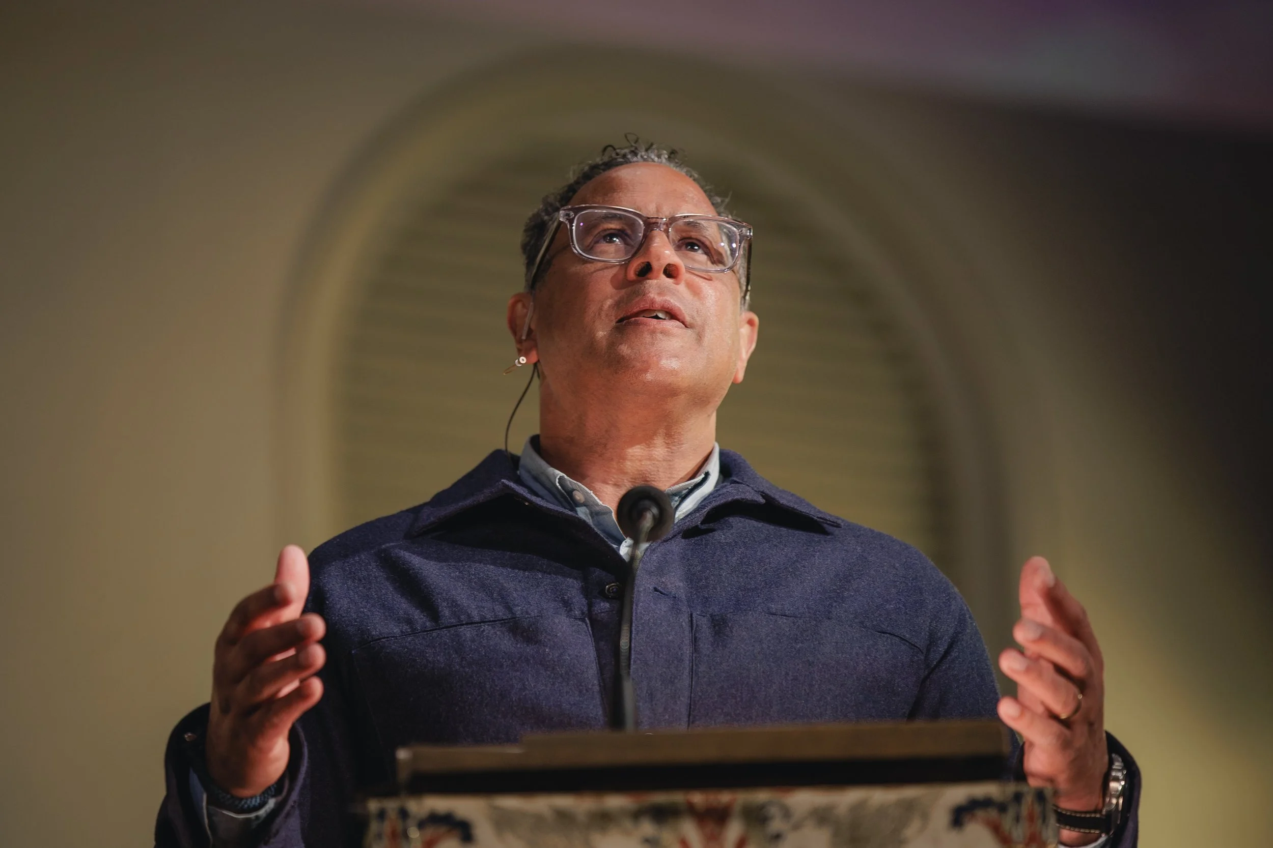 A man wearing glasses is speaking at a podium with a colorful Persian-style rug on its front, gesturing with his hands.