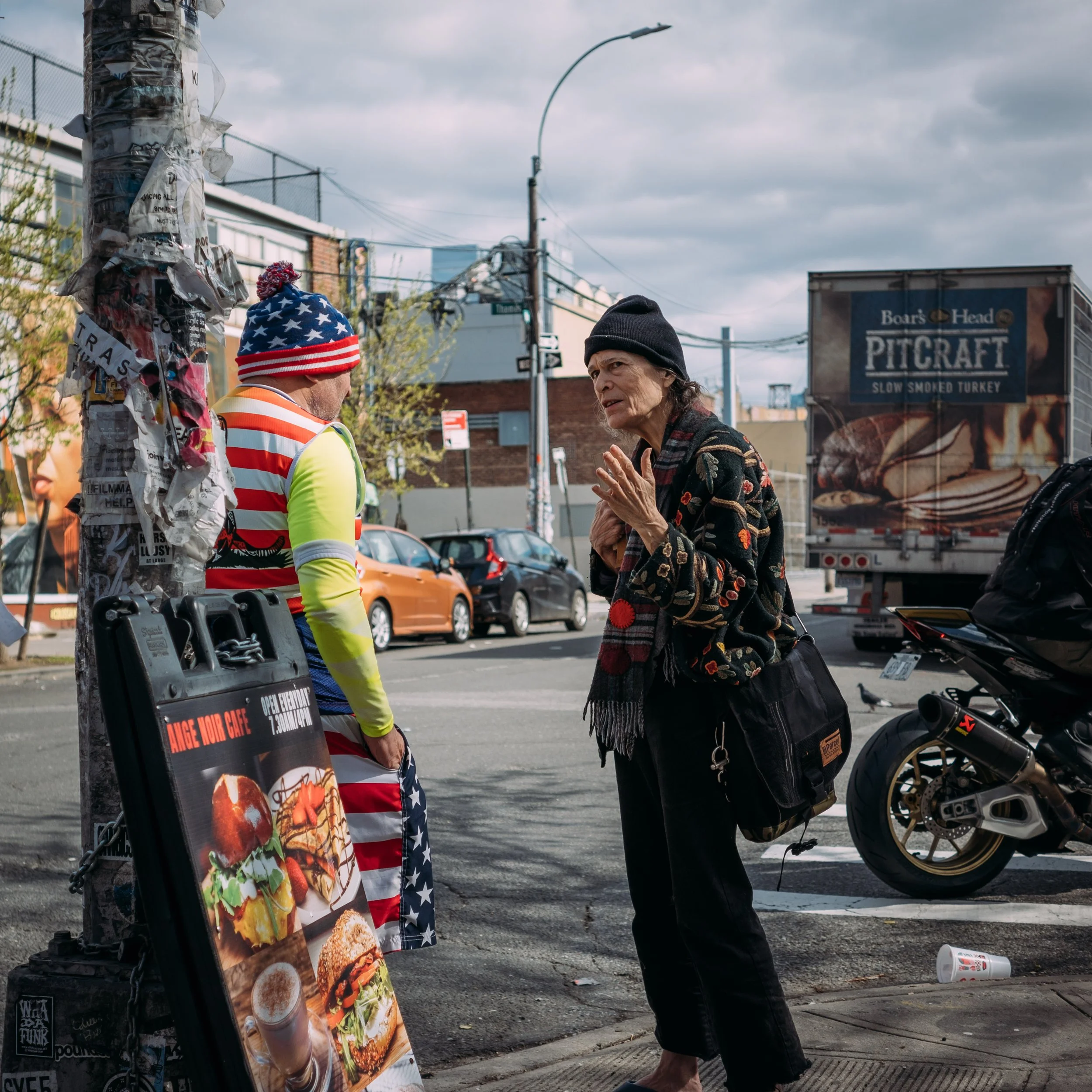 Two older women talking on a city street. One wears a black beanie and a floral coat, and the other has a red, white, and blue knit hat and a neon yellow shirt with an American flag design. A street lamp, a billboard advertising slow smoked turkey, a