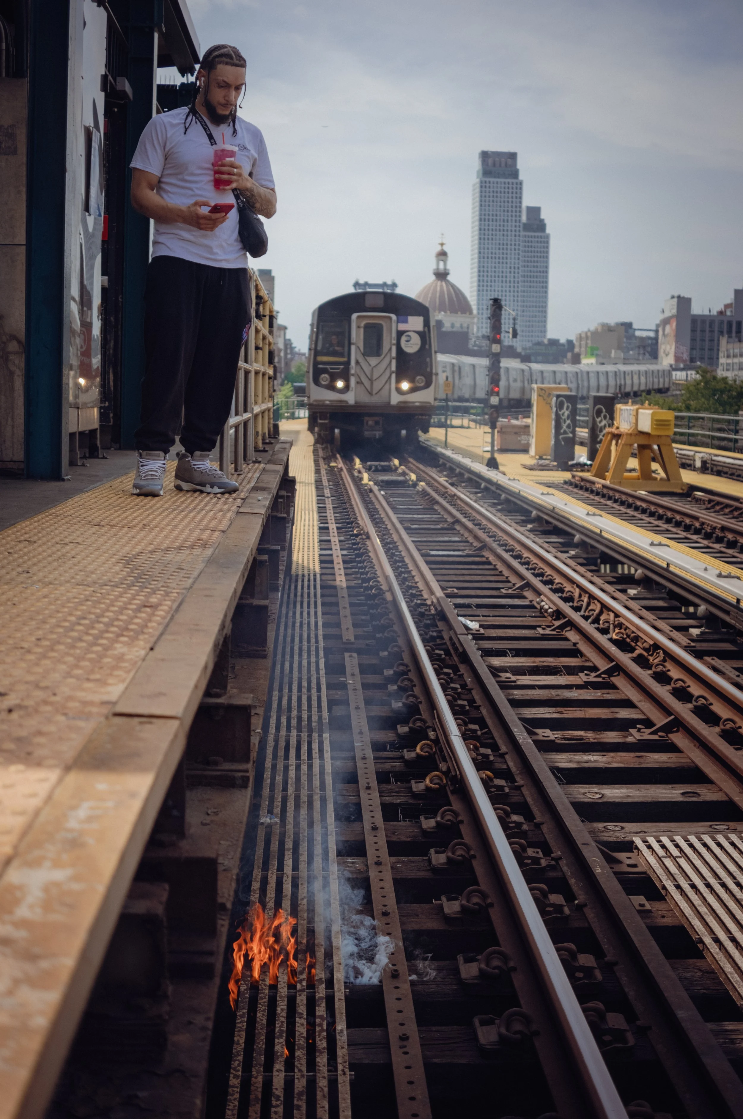 A person standing on a train platform holding a pink beverage and looking at their phone, with a train approaching in the background.