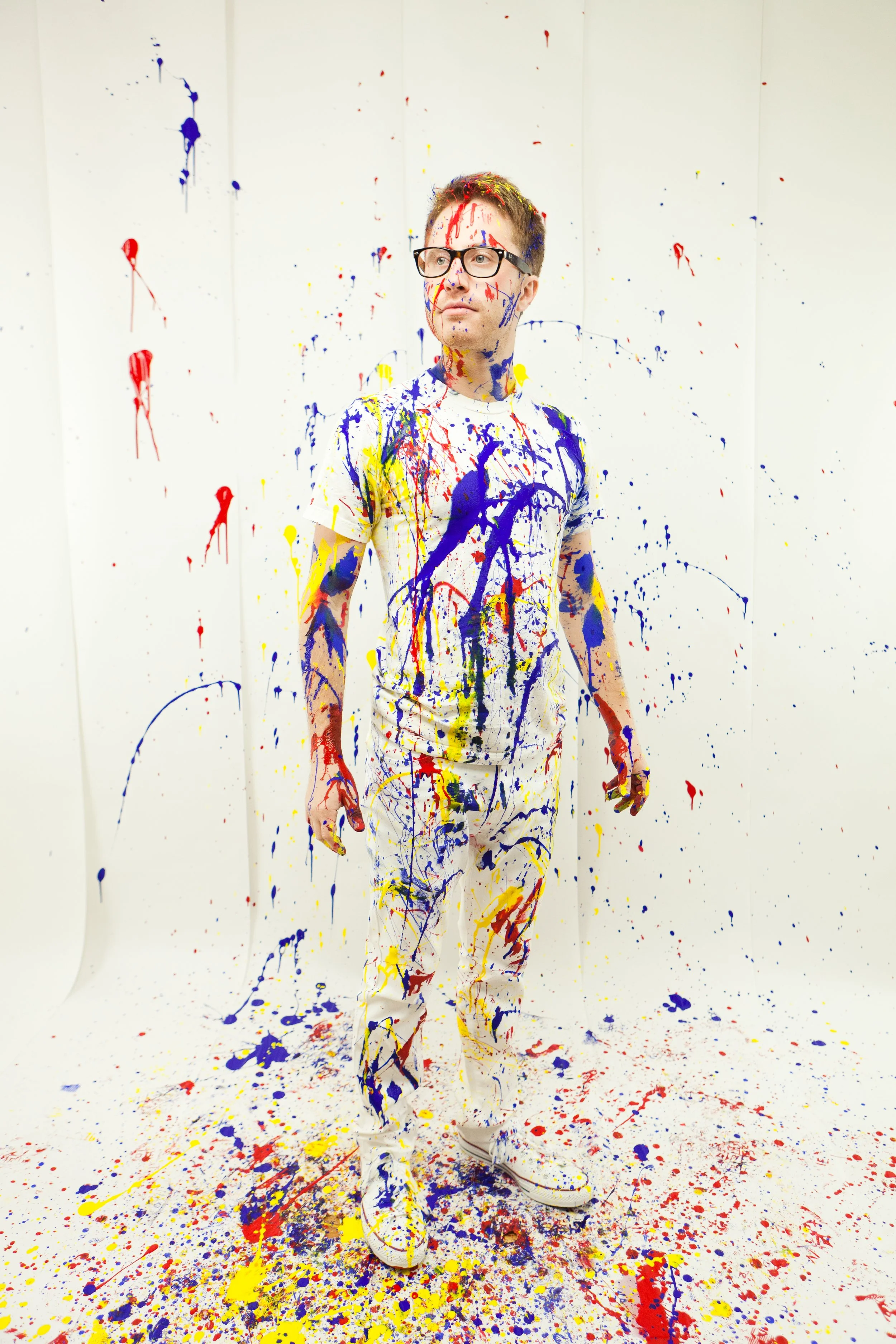 Man wearing glasses in a white t-shirt and white pants, standing in a corner painted with splashes of red, yellow, blue, and purple paint. The wall and floor are covered in similar splatters of paint.