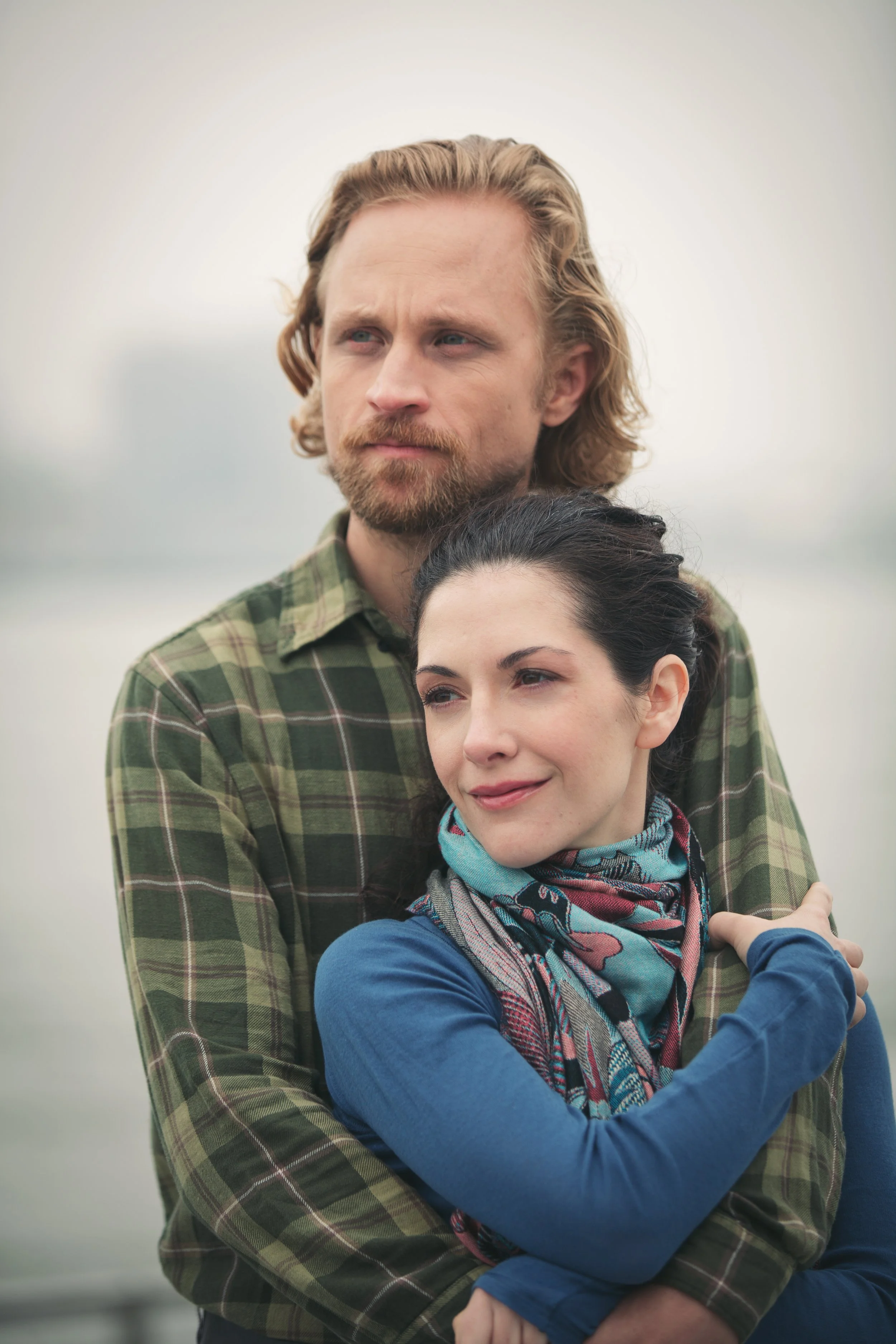 A man with long, wavy blond hair and a beard, wearing a green plaid shirt, standing outdoors with a woman with dark hair and a colorful scarf, smiling and hugging him, against a blurred background.