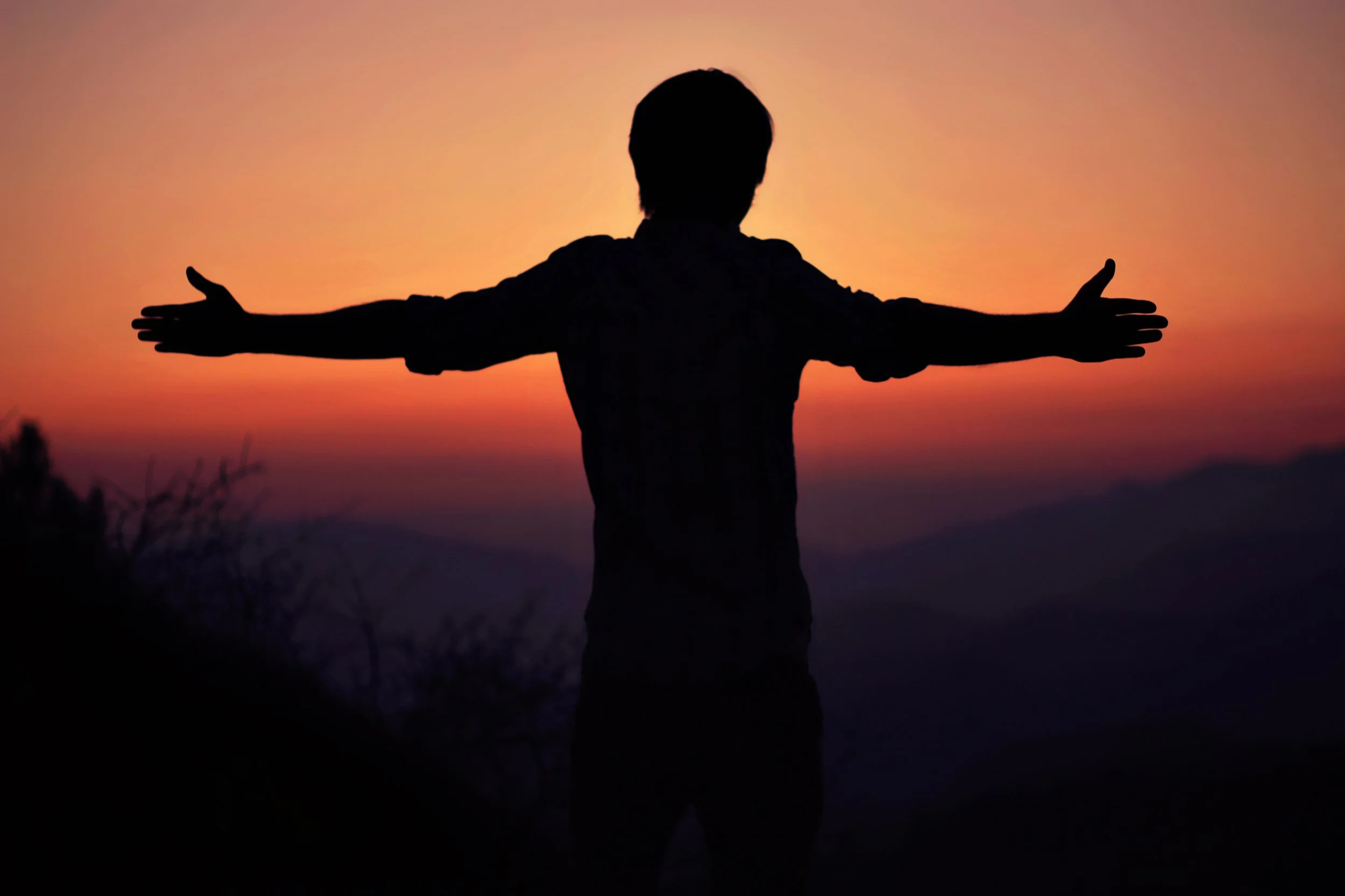 Silhouette of a person with outstretched arms during a sunset or sunrise, with a colorful sky in shades of orange, pink, and purple.