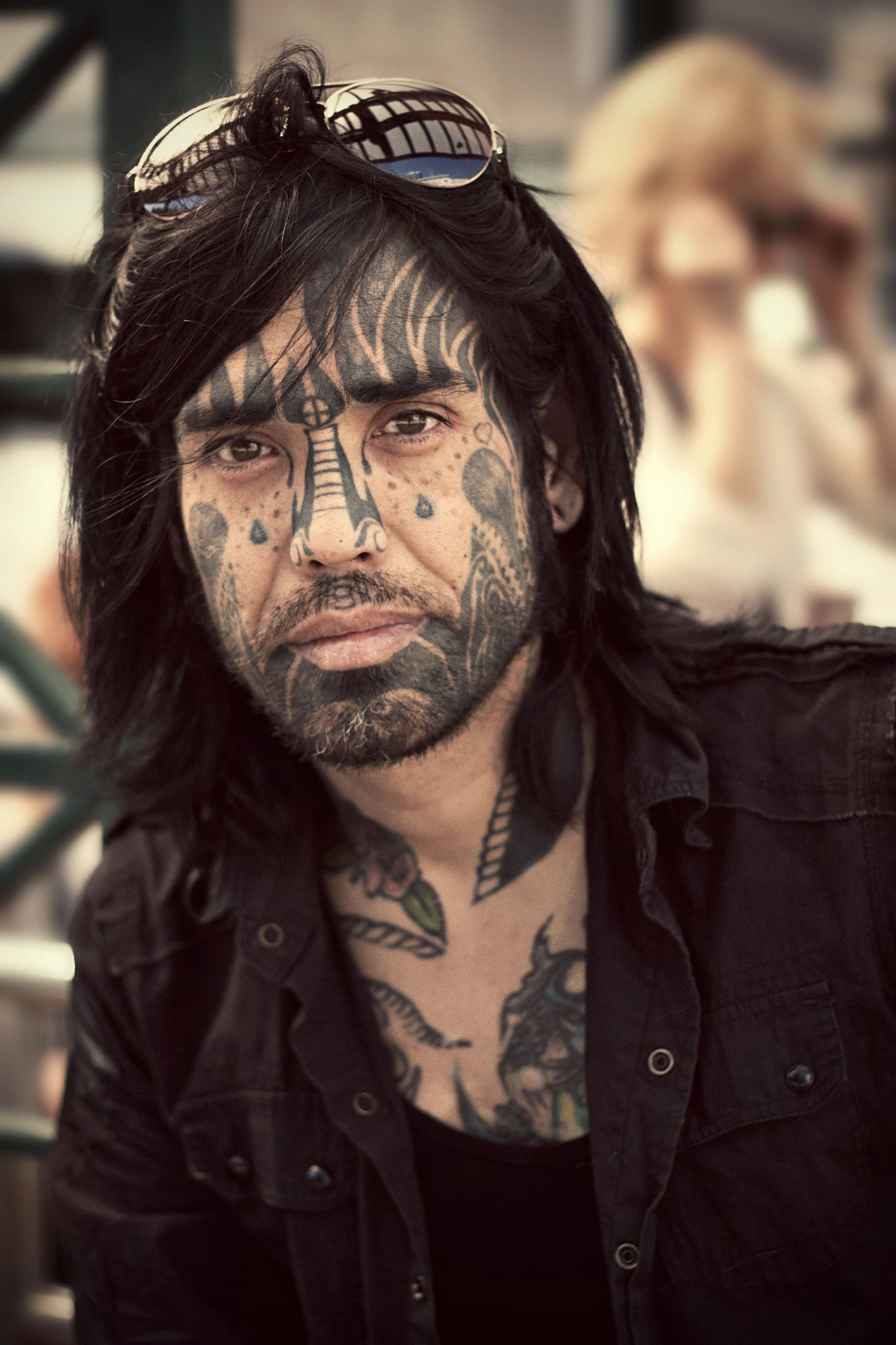 A man with long black hair, tattoos on his face and neck, and sunglasses on his head, looking at the camera. He appears to be in an outdoor or public setting.