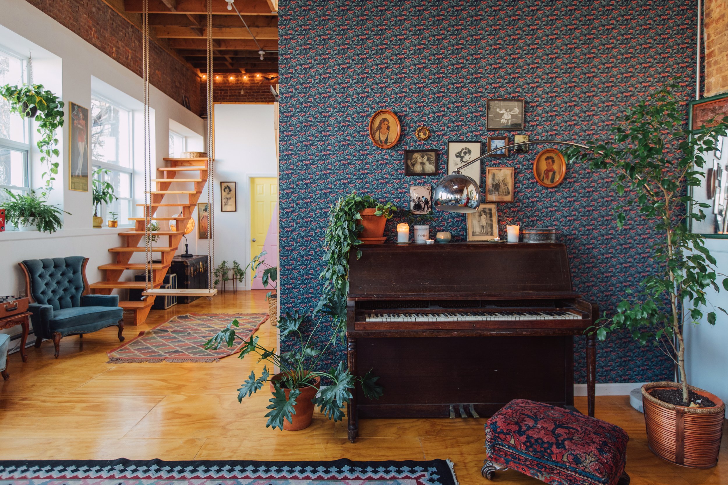 Interior of a vintage-style home with wooden floors, a blue velvet armchair, potted plants, and a wall decorated with framed artwork, candles, and a piano.