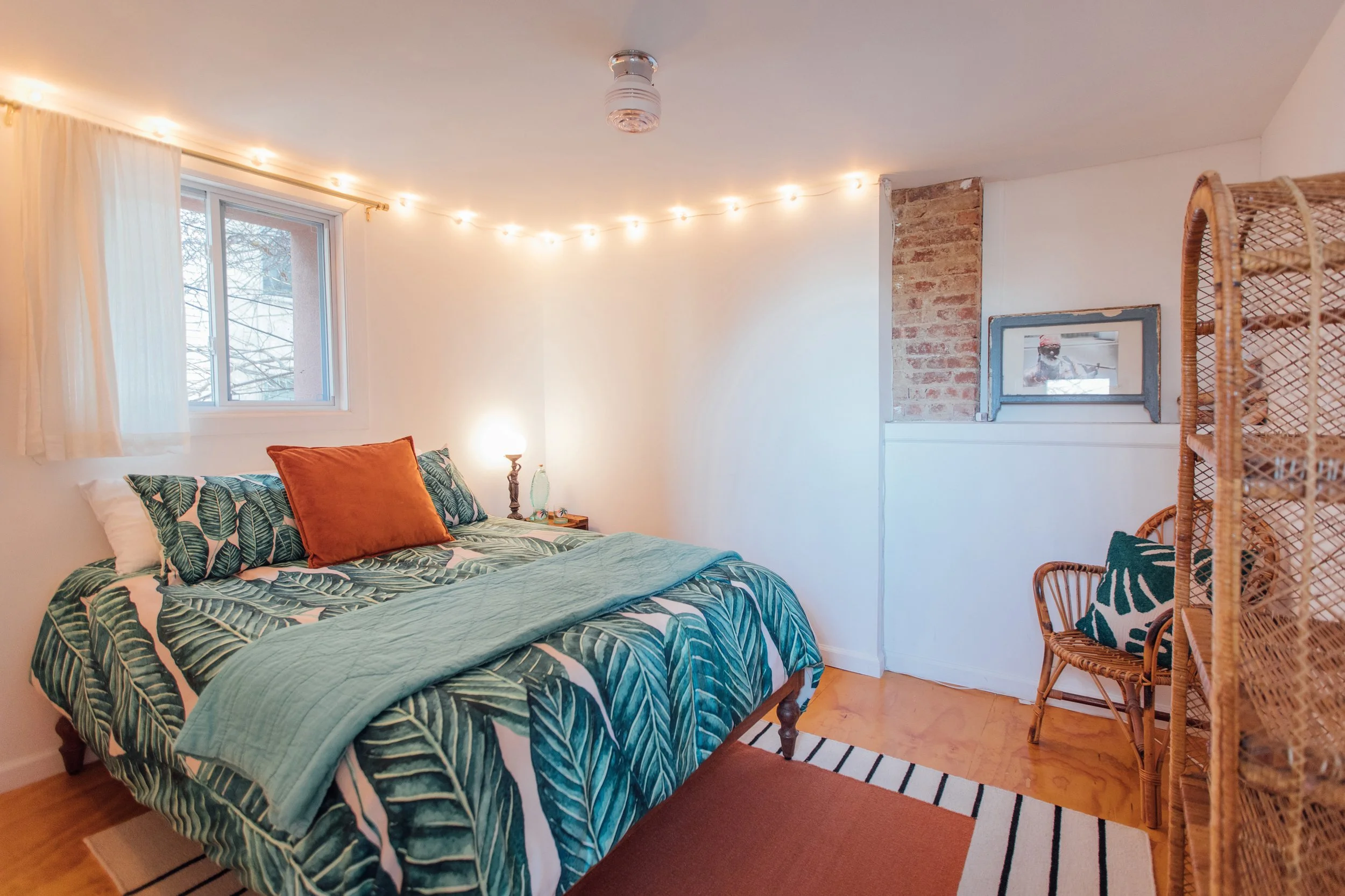 A bedroom with a bed covered in tropical leaf patterned bedding and an orange pillow. There are string lights on the wall, a window with white curtains, a small side table with a lamp, and a wicker chair. Exposed brick accent wall.