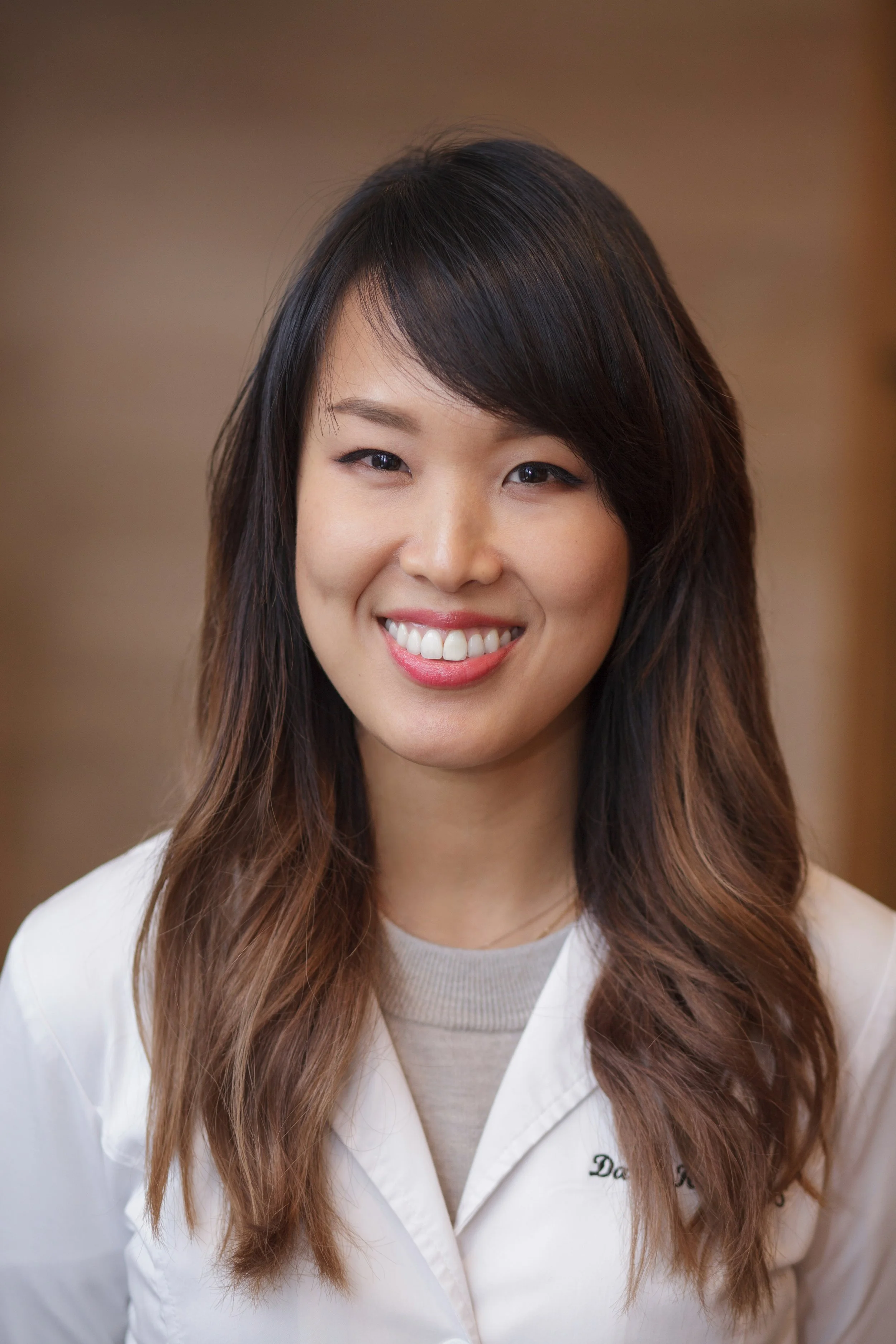 A young woman with medium-length dark brown hair and light brown highlights, smiling and wearing a white coat with embroidered text, in a neutral indoor setting.