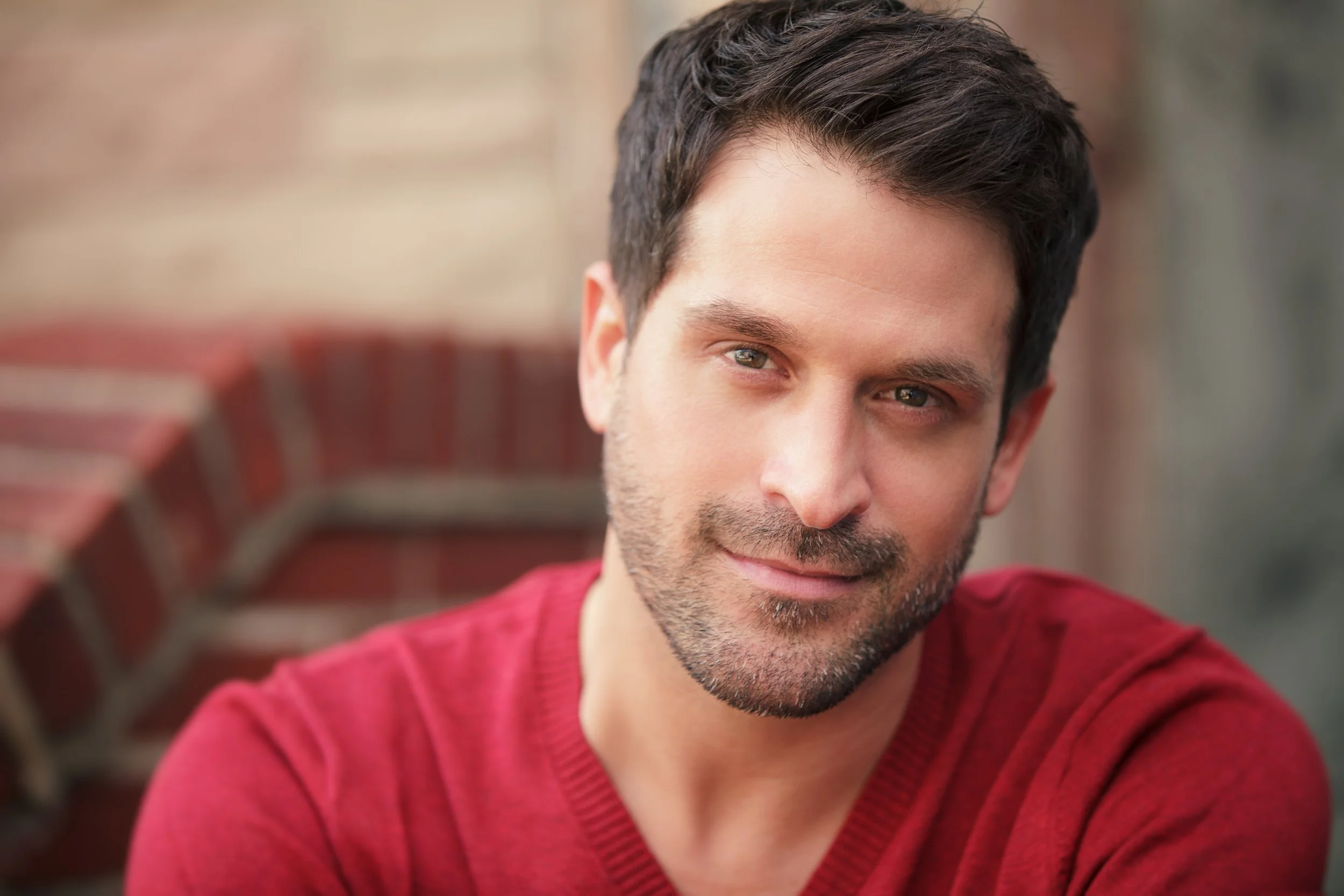 Close-up of a man with dark hair and a beard, wearing a red sweater, smiling slightly, in front of a blurred outdoor background.